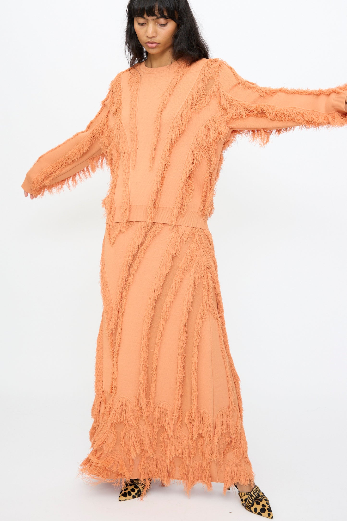 A woman poses with arms outstretched against a white background, wearing the Ulla Johnson Aubree Knit Pullover in Tulip with a matching skirt and leopard print shoes. The peach sweater features jacquard knit and fringe detailing.