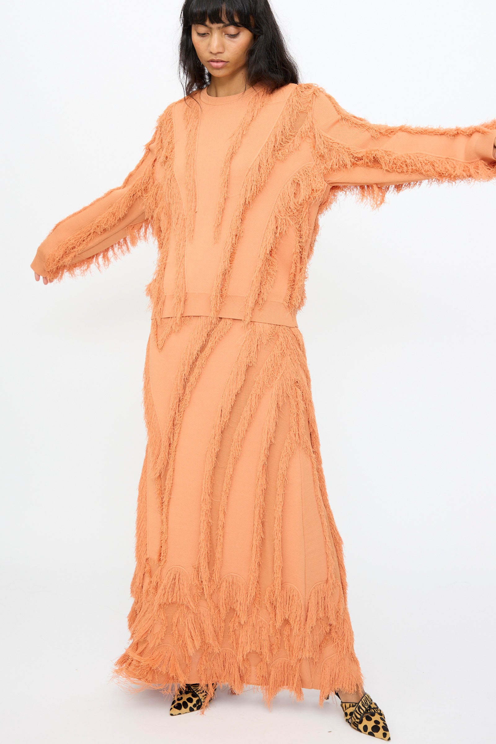 A woman poses with arms outstretched against a white background, wearing the Ulla Johnson Aubree Knit Pullover in Tulip with a matching skirt and leopard print shoes. The peach sweater features jacquard knit and fringe detailing.