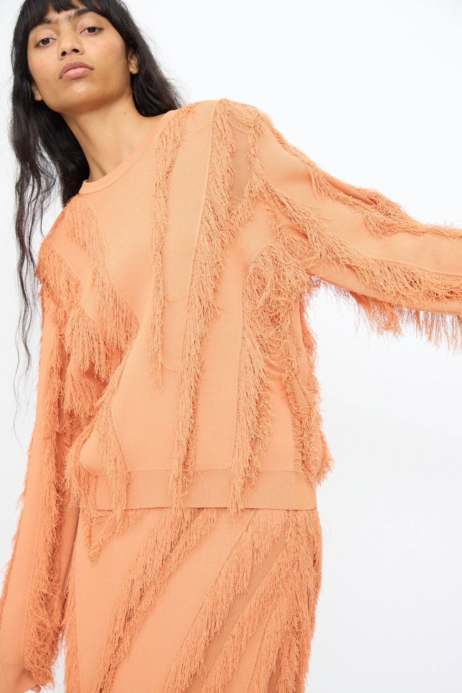A person wearing the Ulla Johnson Aubree Knit Pullover in Tulip, a peach-colored, long-sleeve sweater with a fringe pattern and matching skirt, stands against a plain white background.