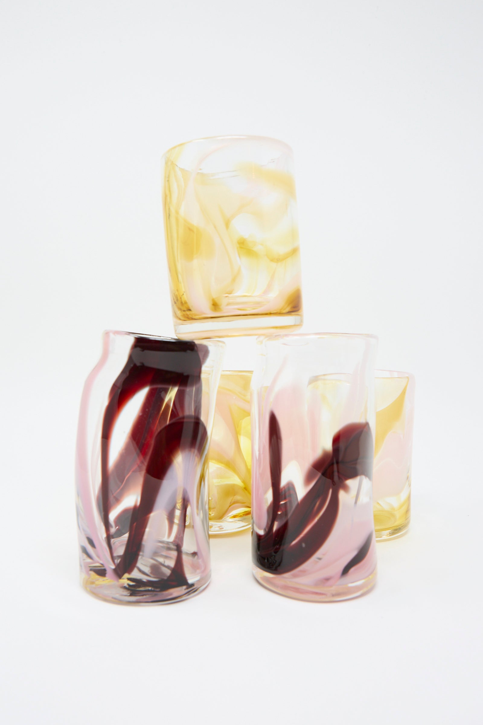 Five artistic Gamay Crinkle Cups by Upstate, featuring handblown glass with swirled patterns in yellow, pink, and dark red hues, are stacked in a pyramid arrangement against a plain background. Each cup showcases an elegant wavy shape that adds a touch of sophistication to any setting.