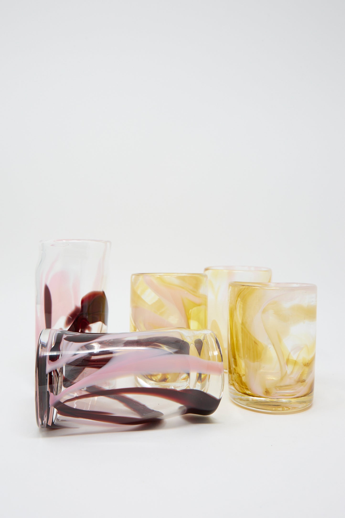 Five Gamay Crinkle Cups by Upstate, featuring handblown designs with swirling patterns in pink, yellow, and white, are arranged on a white surface. One cup is tilted on its side.