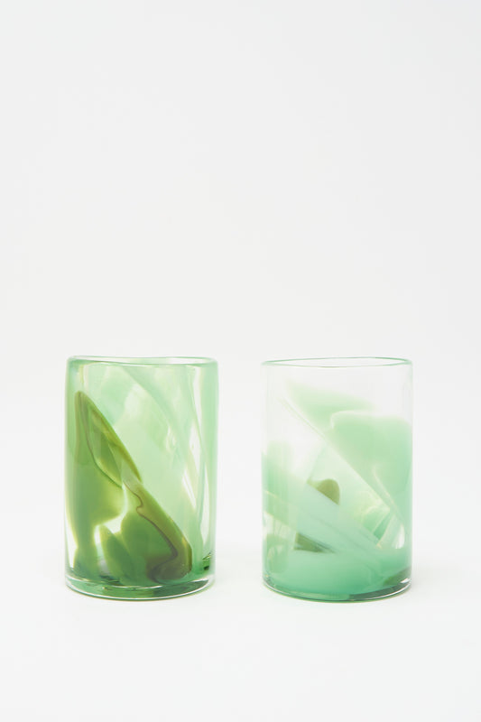 Two Hand Dyed Moss Glass tumblers by Gone, featuring green-tinted, abstract swirled patterns and handmade in NYC, are displayed against a plain white background.