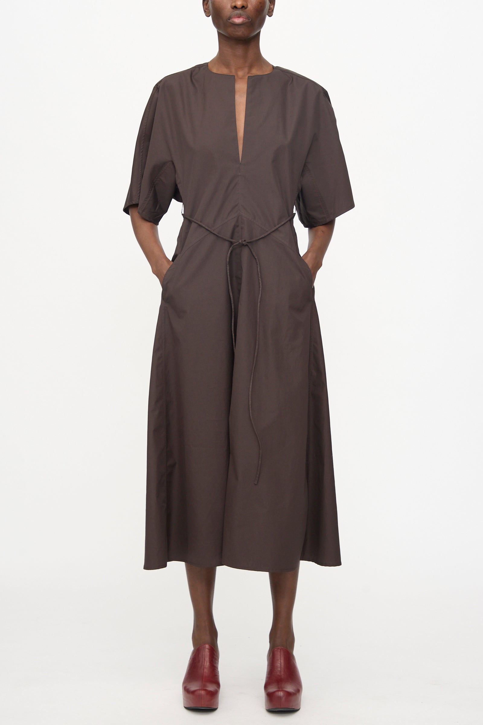A person stands against a white background wearing the Veronique Leroy Belted Kimono Dress in Choco, a relaxed-fit, short-sleeve cotton dress with a deep V-neckline and waist tie, paired with red heeled shoes.