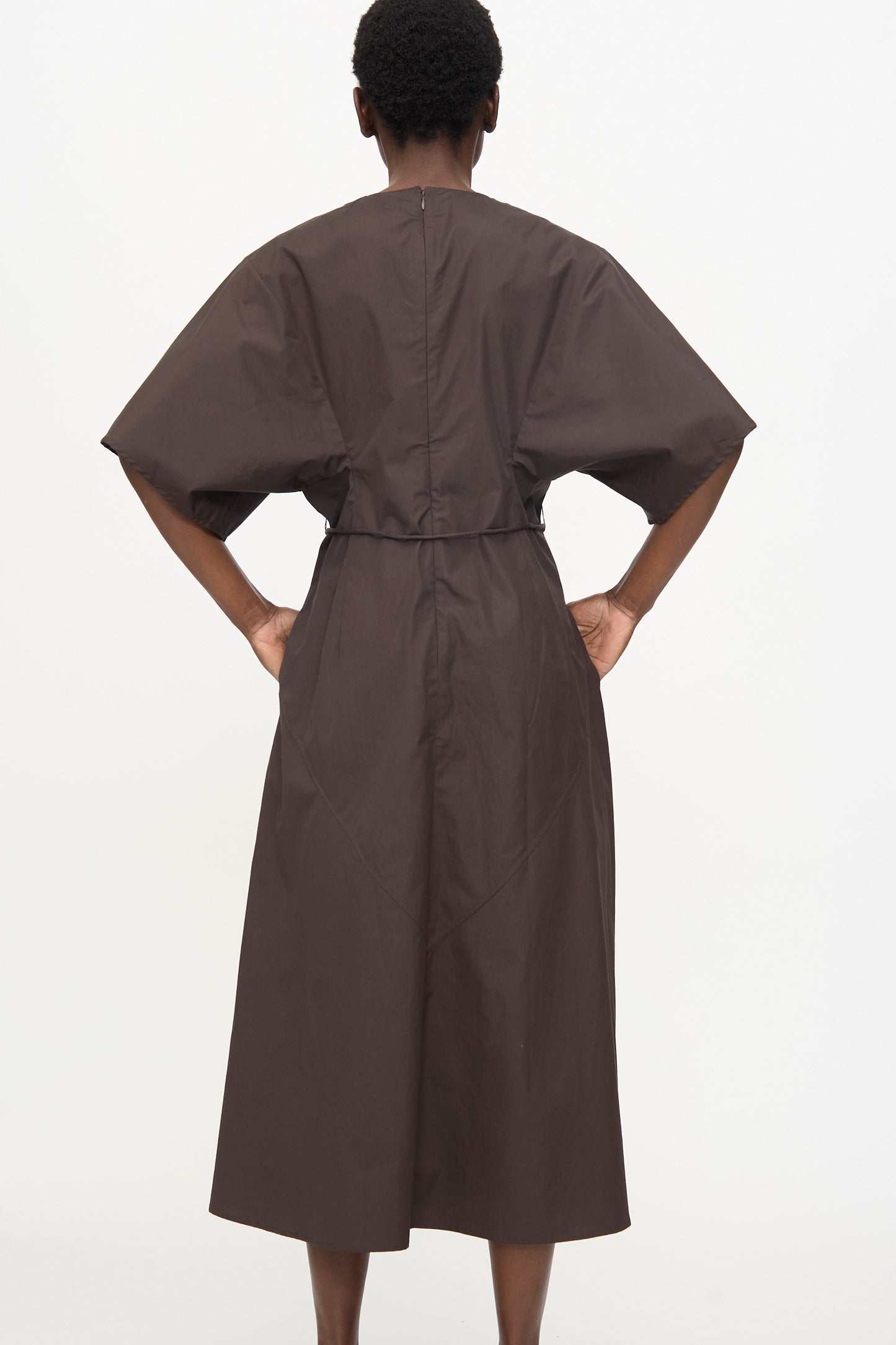 Person stands with hands on hips, wearing the Veronique Leroy Belted Kimono Dress in Choco—a dark brown, relaxed-fit, short-sleeved midi dress—photographed from the back against a plain white background.