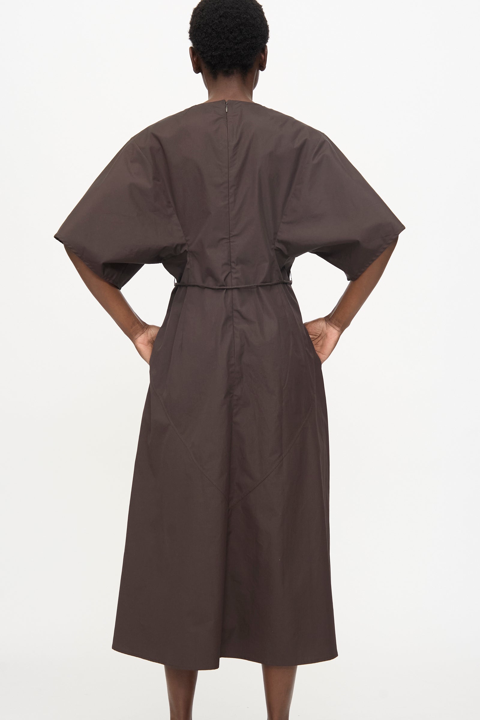 Person stands with hands on hips, wearing the Veronique Leroy Belted Kimono Dress in Choco—a dark brown, relaxed-fit, short-sleeved midi dress—photographed from the back against a plain white background.