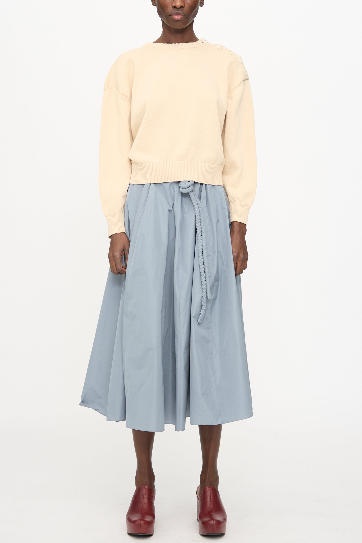 A person wearing the Veronique Leroy Button Shoulder Boxy Sweater in Cream, a light blue tied-waist midi skirt, and red heeled shoes stands against a plain white background.