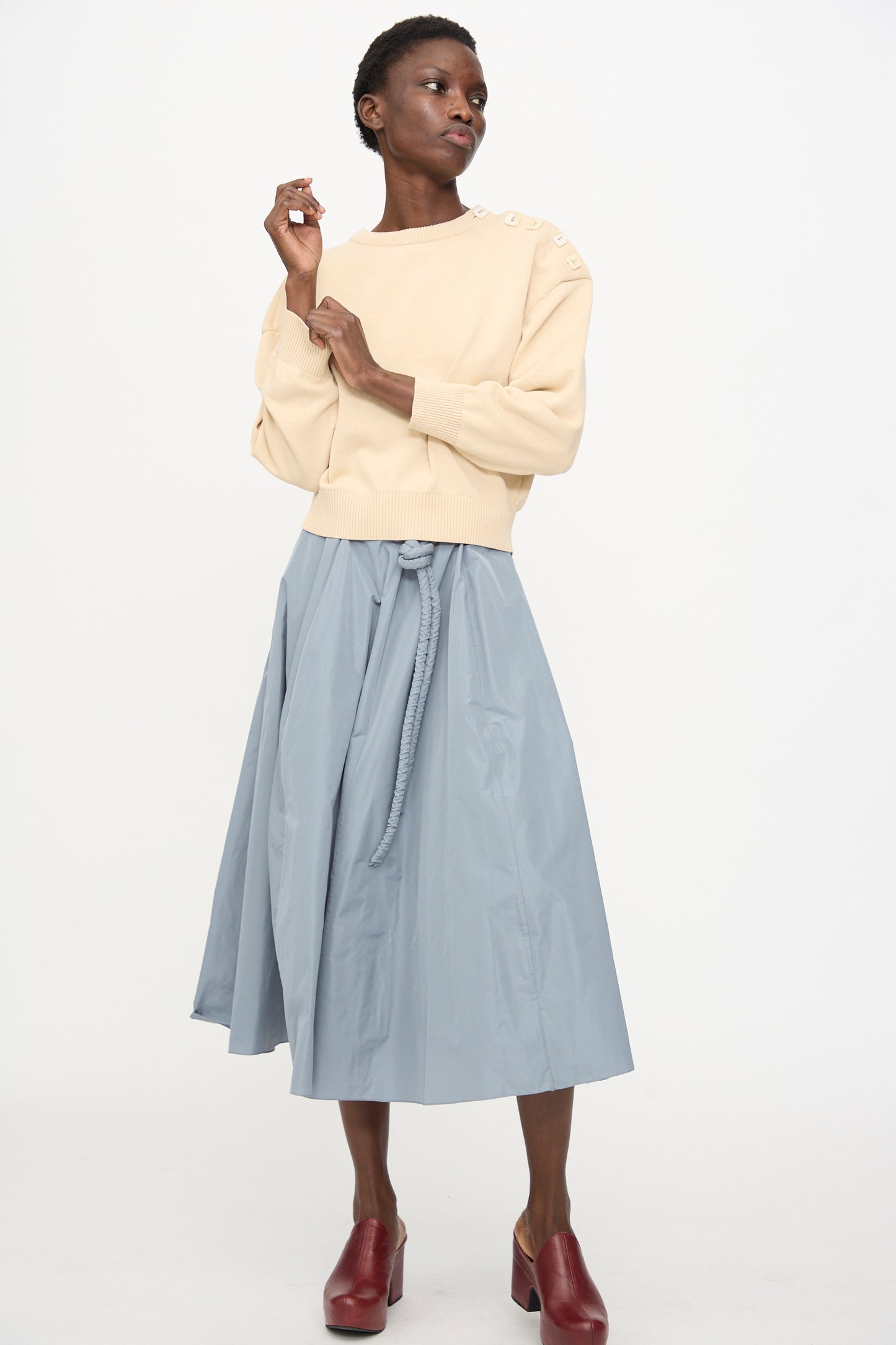A person wears the Veronique Leroy Button Shoulder Boxy Sweater in Cream, styled with a high-waisted light blue midi skirt and burgundy heeled mules, standing against a plain white background.