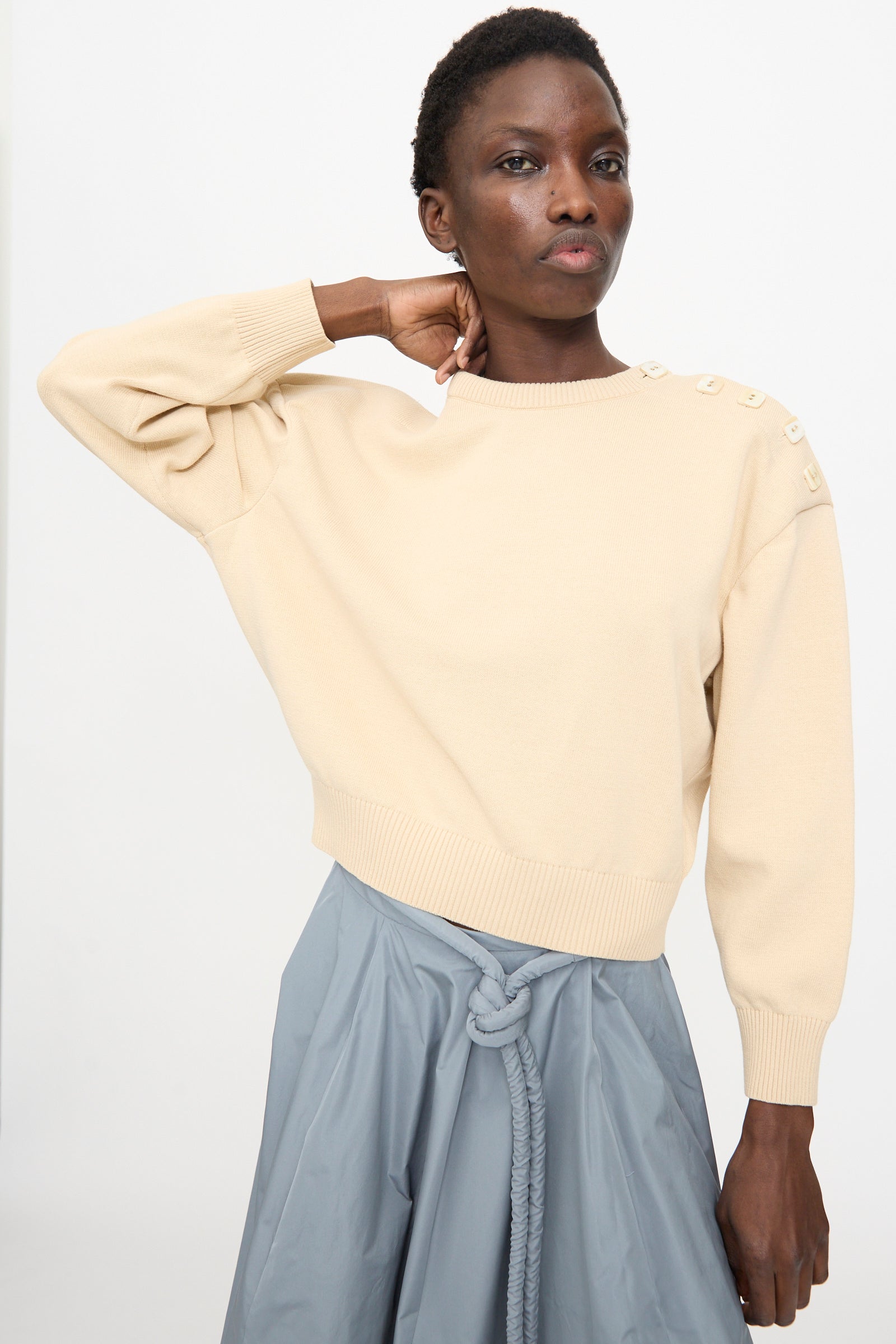 A person wears the Veronique Leroy Button Shoulder Boxy Sweater in Cream, styled with a blue skirt and rope belt, posing against a plain light background.