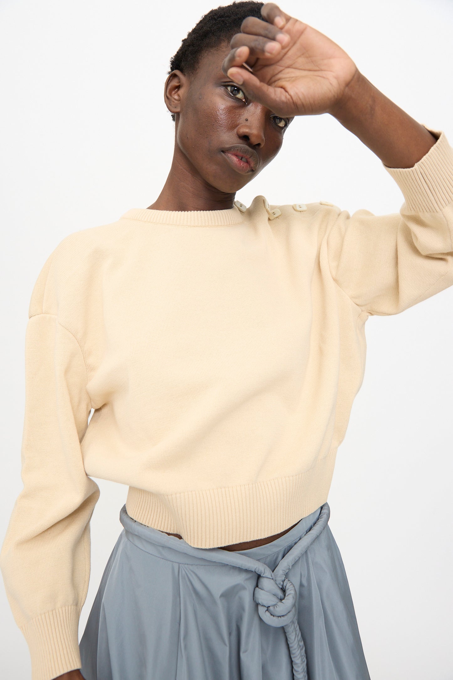 Wearing the Veronique Leroy Button Shoulder Boxy Sweater in Cream, a person stands against a plain background with one arm raised to partially cover their face.