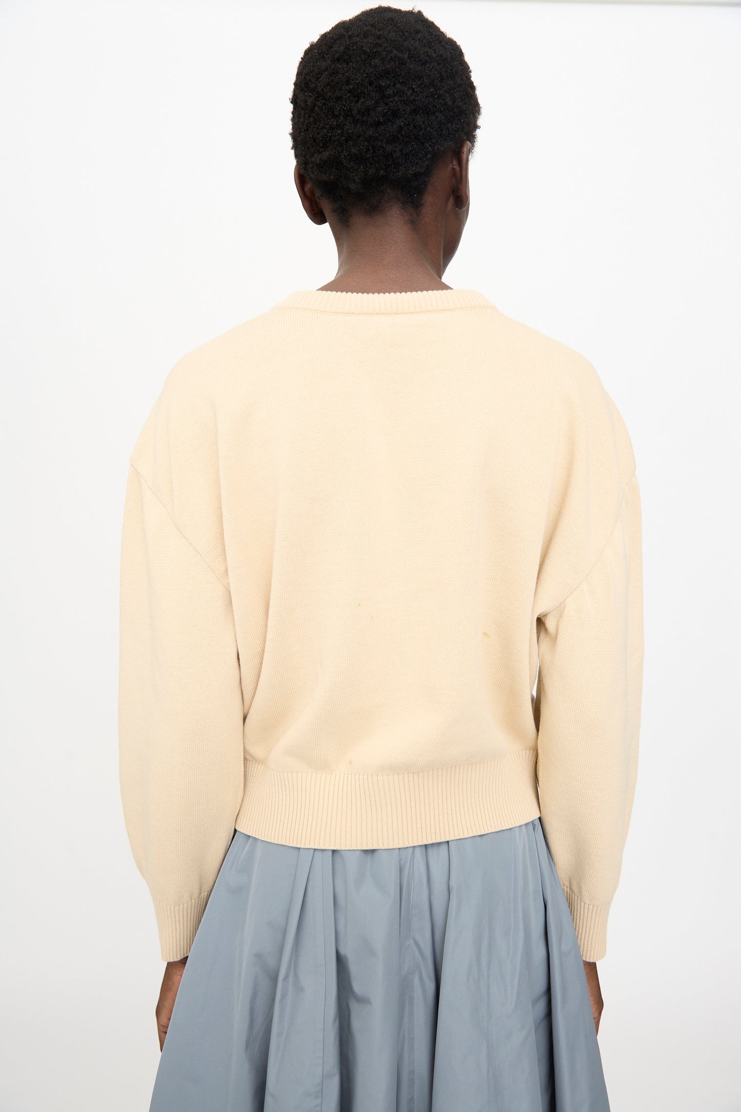 A person stands with their back to the camera, wearing the Veronique Leroy Button Shoulder Boxy Sweater in Cream paired with a light blue skirt, set against a plain background.