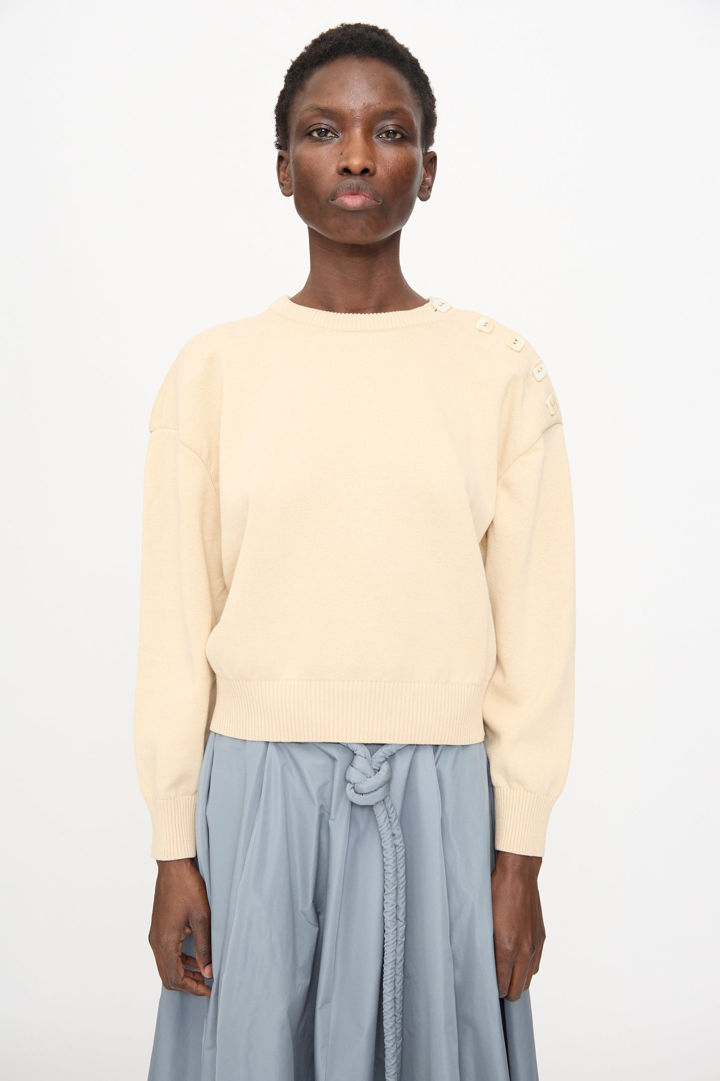 A person wears the Veronique Leroy Button Shoulder Boxy Sweater in Cream, styled with a light blue skirt featuring a knotted belt, standing against a plain background.