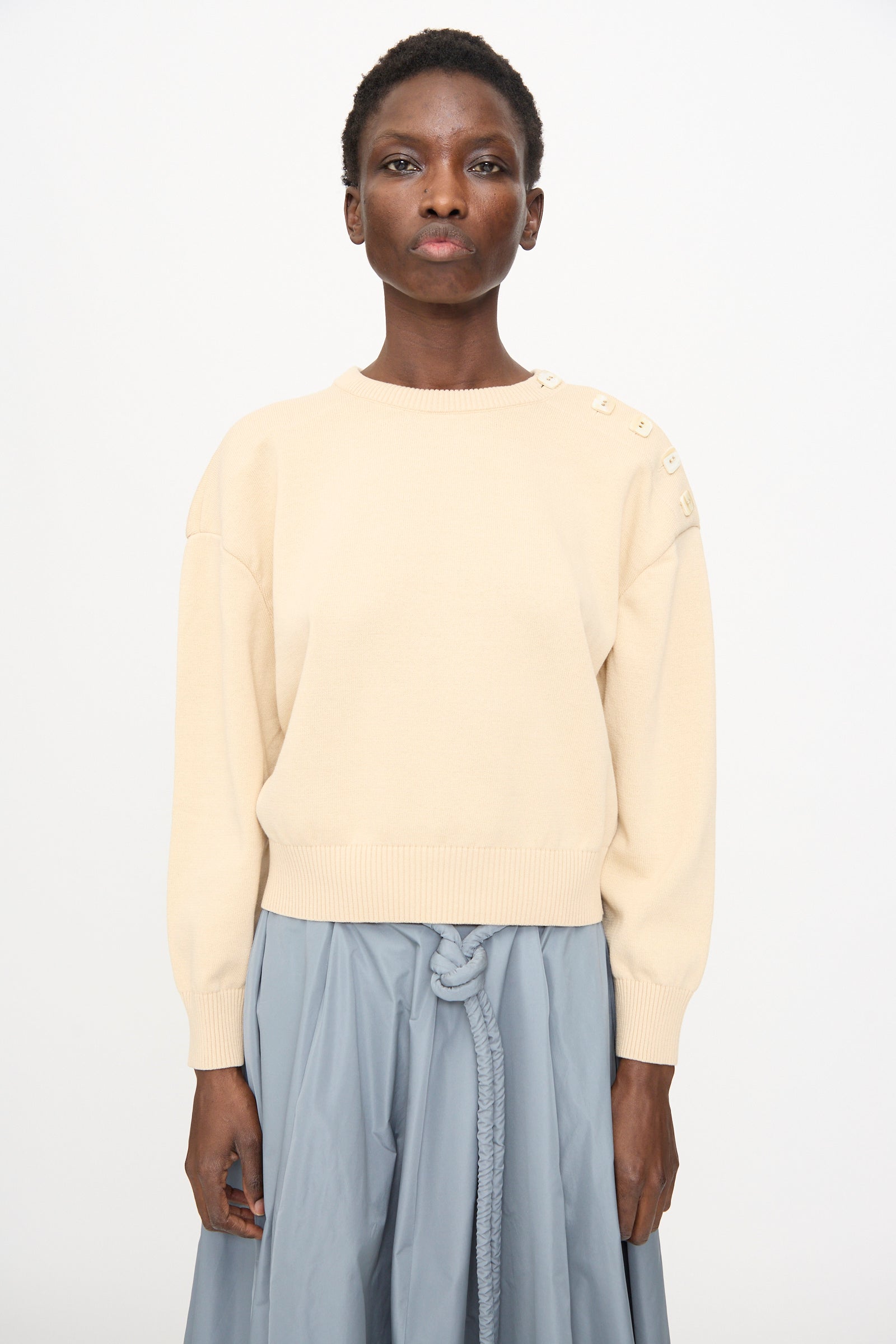 A person wears the Veronique Leroy Button Shoulder Boxy Sweater in Cream, styled with a light blue skirt featuring a knotted belt, standing against a plain background.