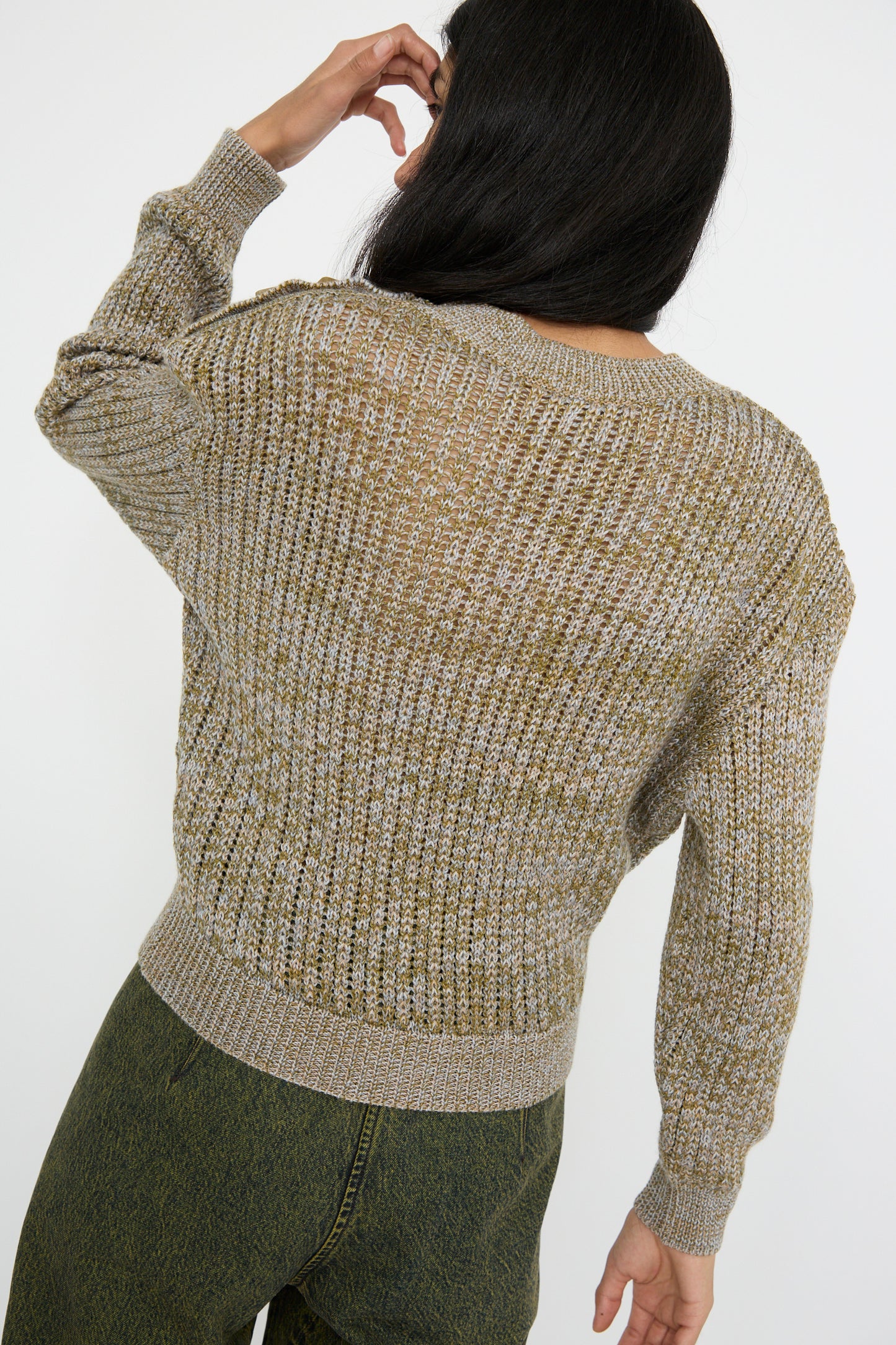 A rear view of a person with long dark hair wearing the Button Shoulder Sweater in Meadown by Veronique Leroy, paired with dark green pants, adjusting their hair with one hand.