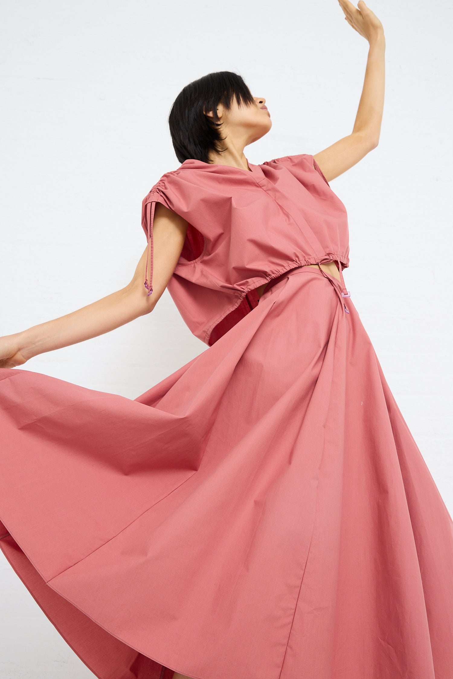 Woman in a Cotton Poplin Gathered Top in Canyon by Veronique Leroy and flowing pink dress with adjustable ruche detail, her arm extended upwards against a white background.