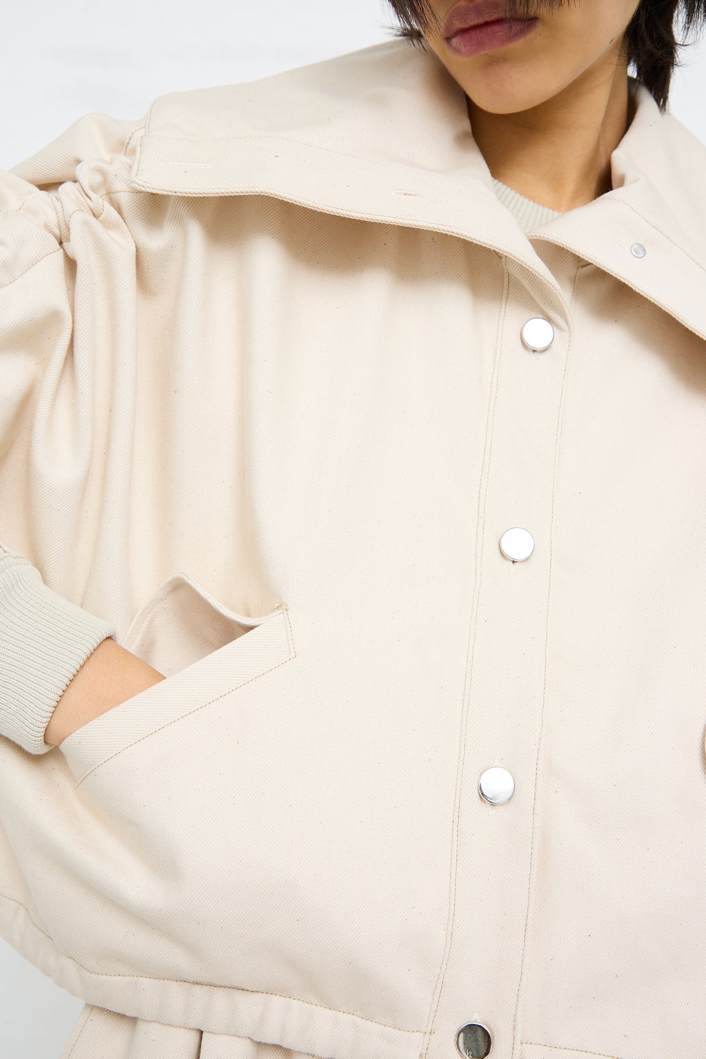 Close-up of a person wearing a beige, short sleeve Veronique Leroy denim high neck jacket in chalk with white snap button closure.
