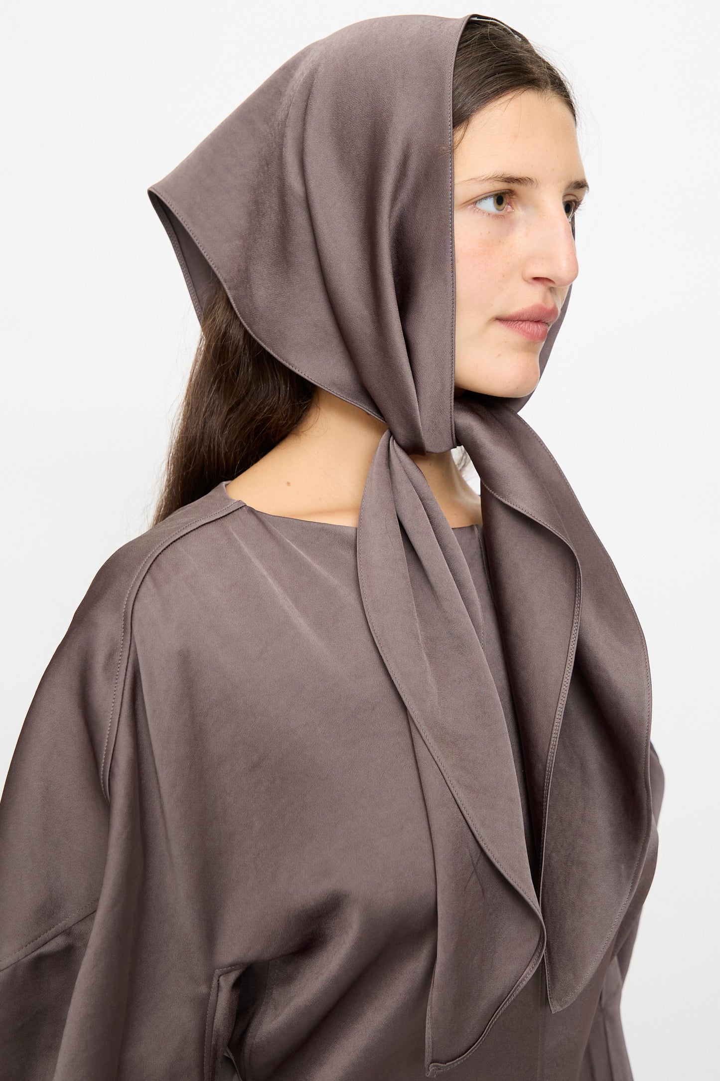 A woman in the Veronique Leroy Dress with Scarf in Wood, featuring a taupe headscarf and sculpted sleeve midi dress with a fitted waist, looks to the side against a plain light background.