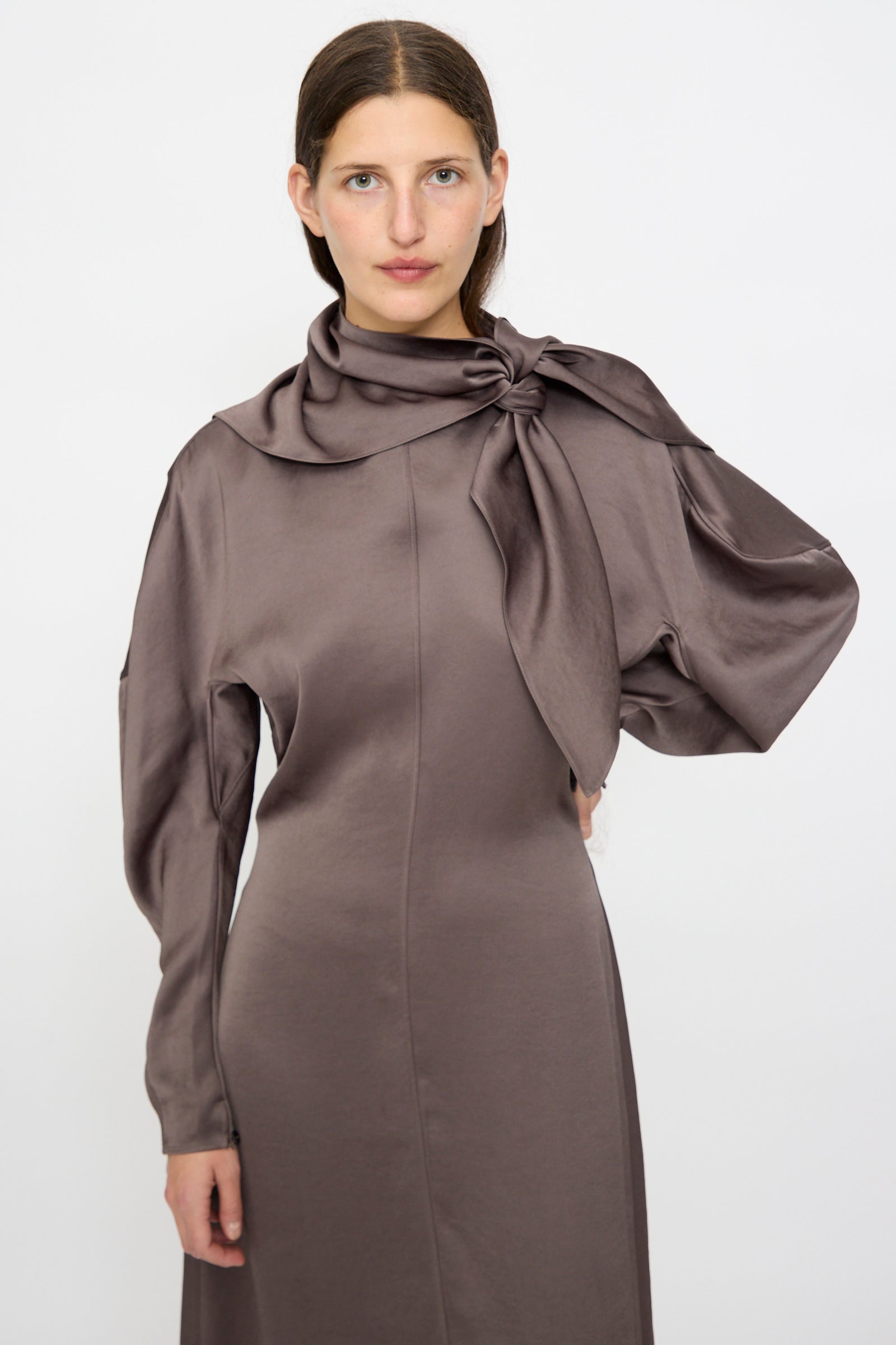 A woman faces forward wearing the Veronique Leroy Dress with Scarf in Wood, featuring long sleeves, a fitted waist, and an oversized knotted shoulder bow that adds elegant structure to her style.