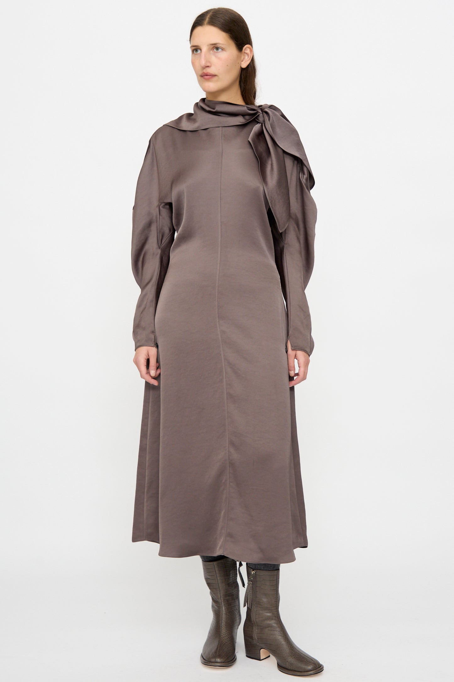 A woman stands against a plain background wearing the Veronique Leroy Dress with Scarf in Wood, featuring a fitted waist, taupe midi length, draped scarf detail, and gray mid-calf boots.