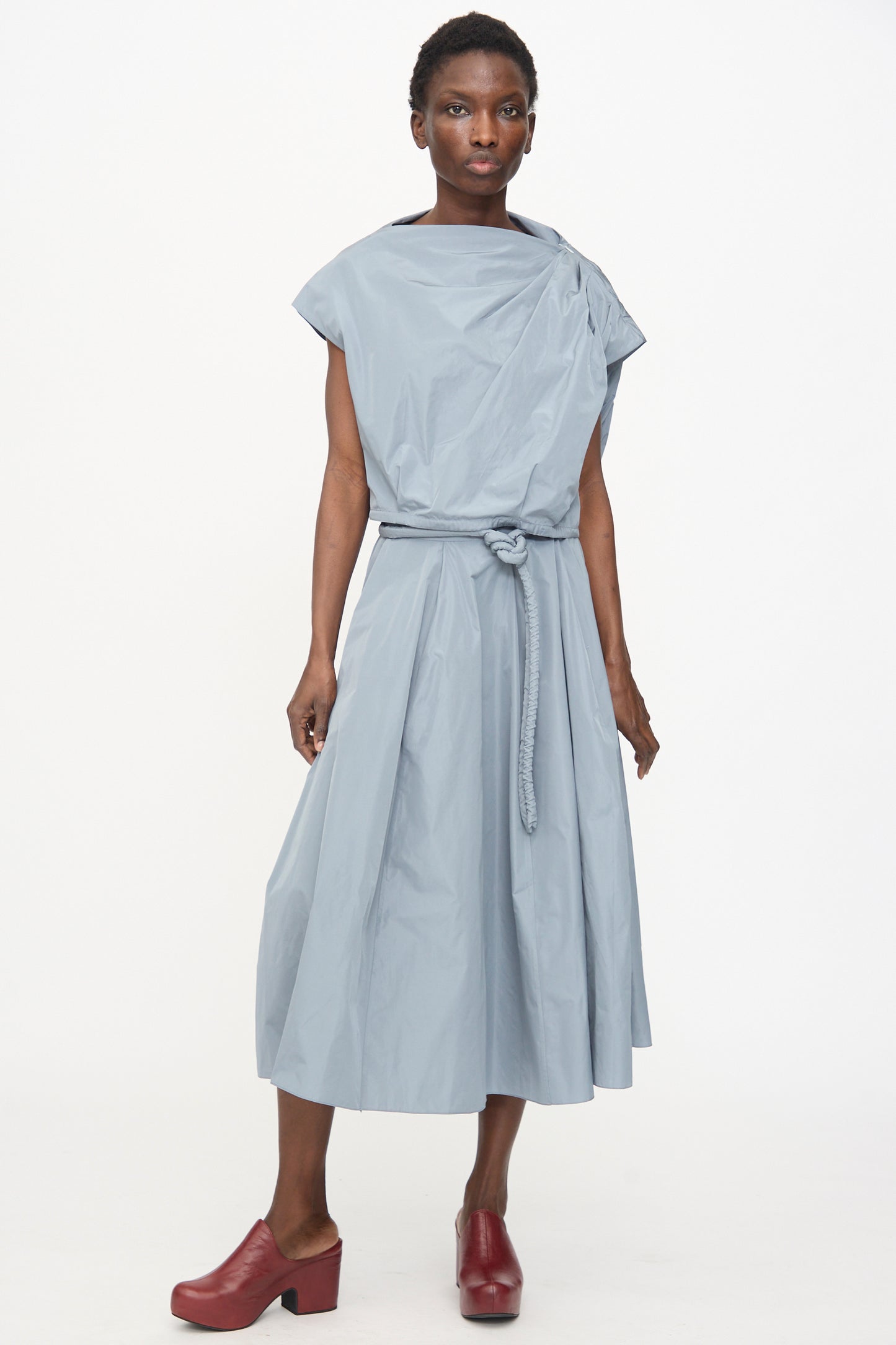 Model wears the Veronique Leroy Gather Shoulder Top in Storm, styled as a belted, sleeveless midi dress with a draped neckline and red platform shoes, standing against a plain white background.