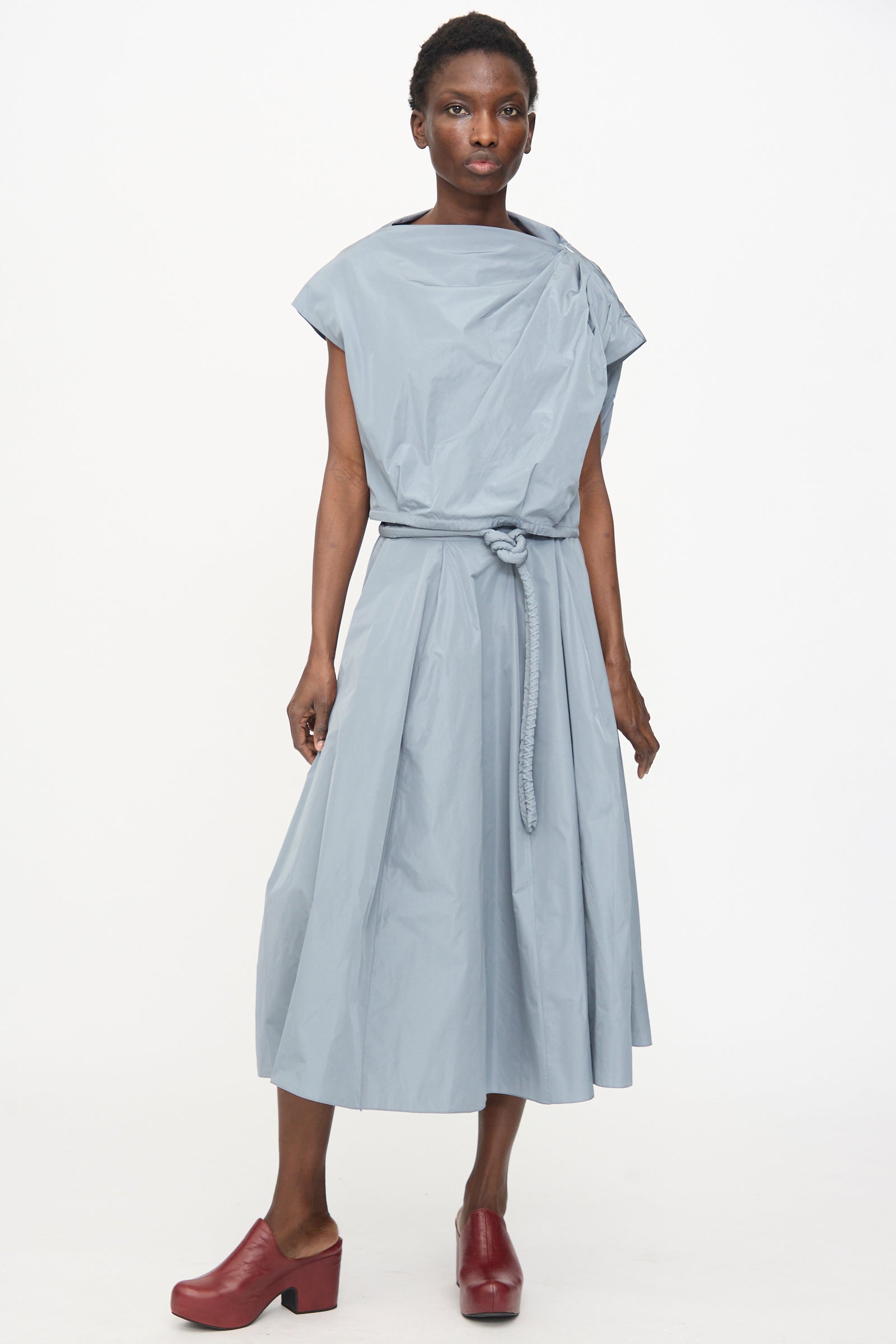 Model wears the Veronique Leroy Gather Shoulder Top in Storm, styled as a belted, sleeveless midi dress with a draped neckline and red platform shoes, standing against a plain white background.
