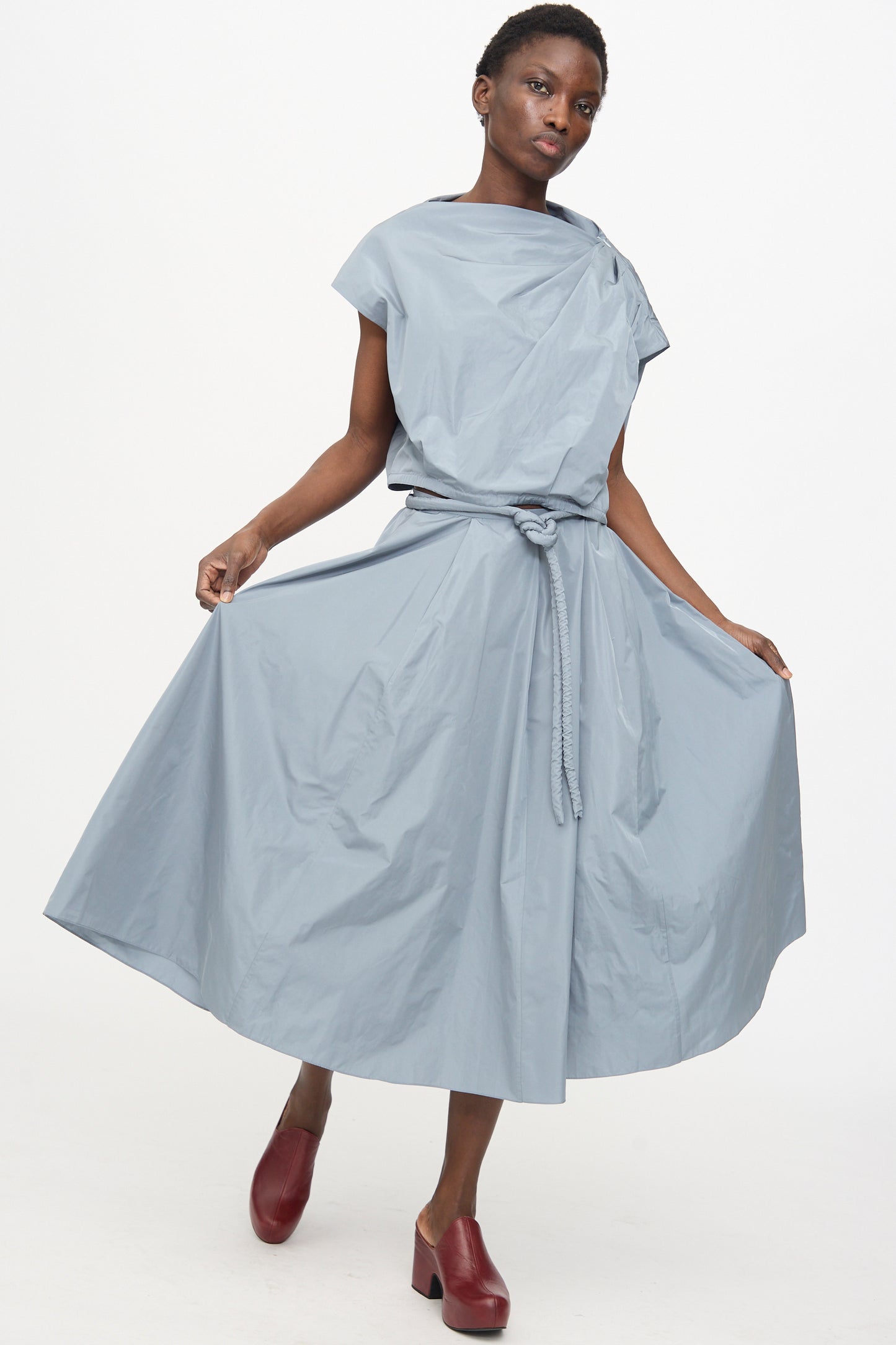 A person wears the Veronique Leroy Gather Shoulder Top in Storm, styled against a white background while holding the sides of their top, and finishes the look with dark red shoes.