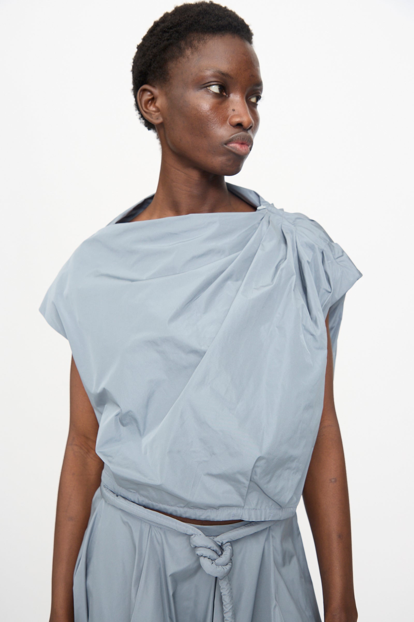 A person wears the Veronique Leroy Gather Shoulder Top in Storm—light gray, sleeveless with an adjustable cinched hem—paired with a matching belted skirt, standing against a plain white background and looking to the side.