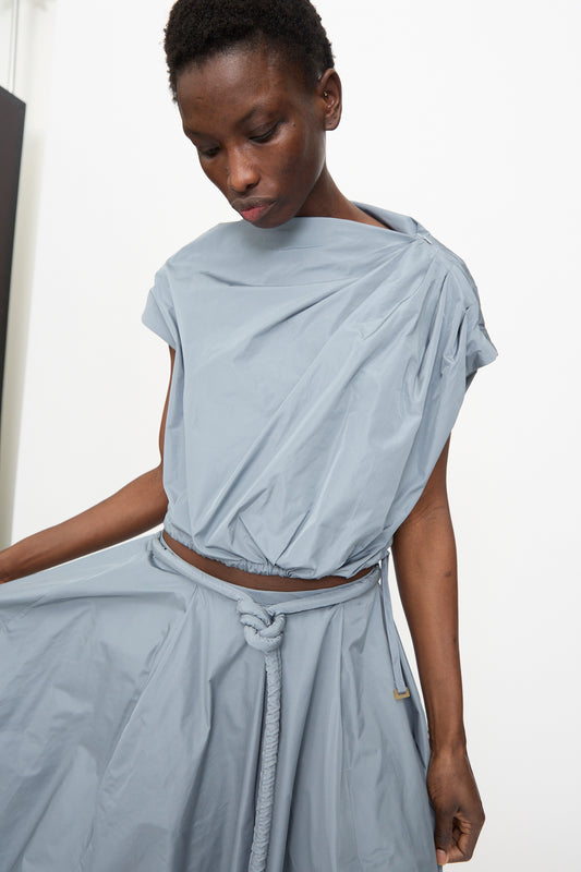 A model wears the Veronique Leroy Gather Shoulder Top in Storm, a light blue taffeta sleeveless piece with an adjustable cinched hem, paired with a matching skirt and knotted rope belt, against a plain background.