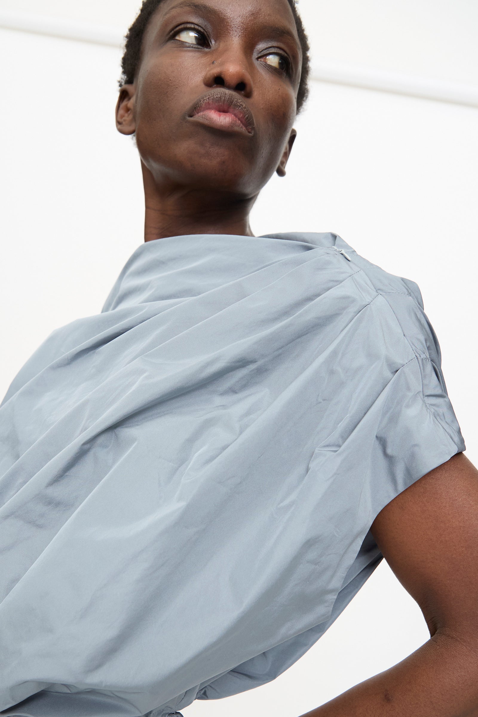 Model wears the Veronique Leroy Gather Shoulder Top in Storm—a light gray, sleeveless top with a cinched adjustable hem—while looking upward and to the side against a plain, light background.