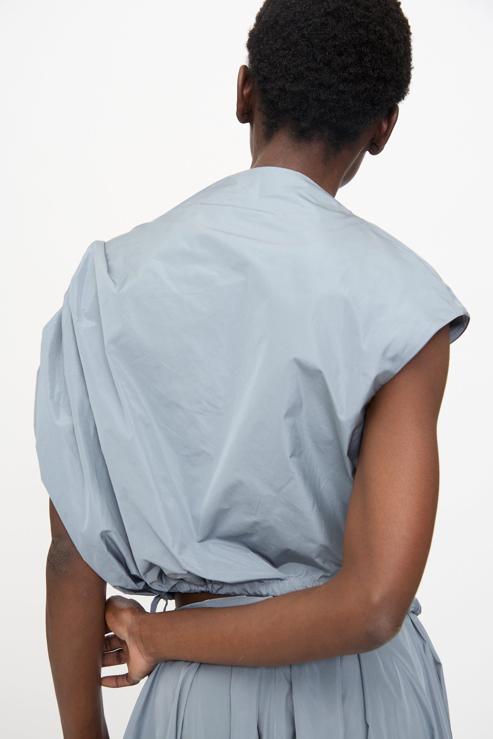 A person with short hair, wearing the Veronique Leroy Gather Shoulder Top in Storm—a light blue taffeta top with an adjustable cinched hem—stands facing away from the camera, holding their waist.