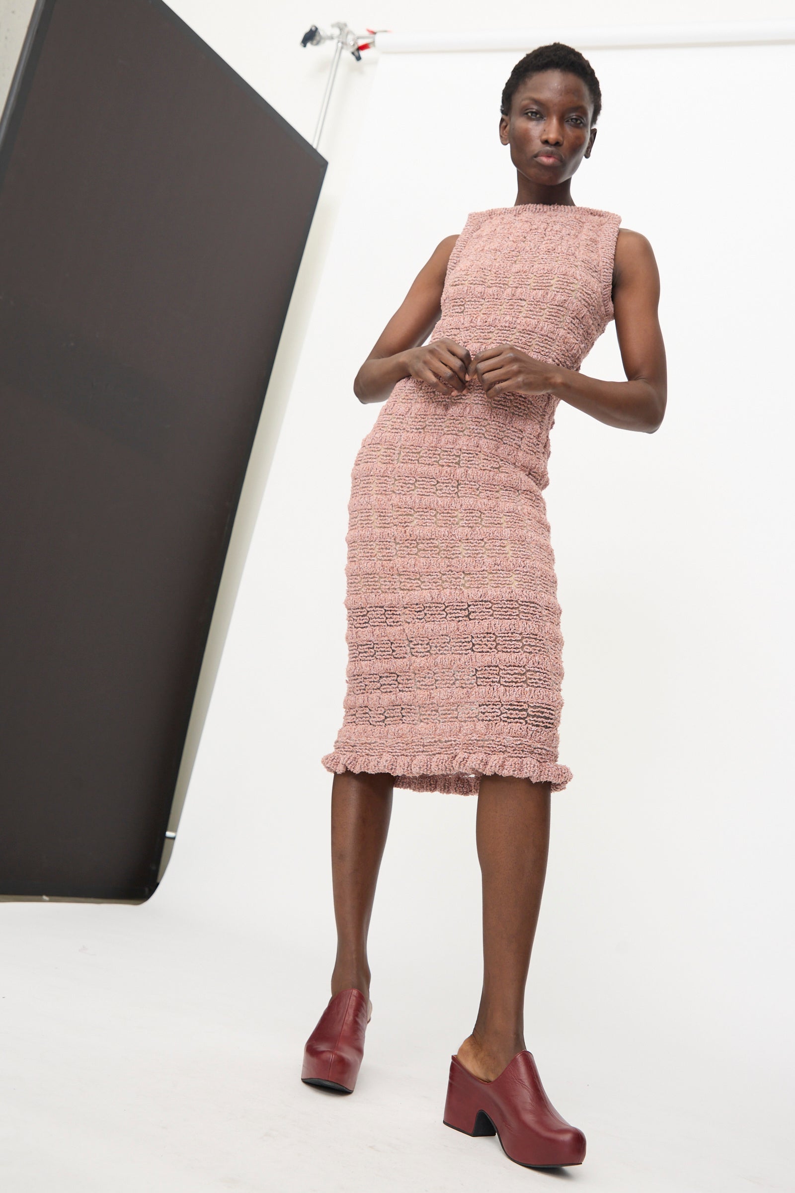 A person poses in a studio with a white backdrop and black panel, wearing the Veronique Leroy Smock Knit Dress in Dusk—a pink sleeveless cotton blend dress with textured details—and red platform shoes.