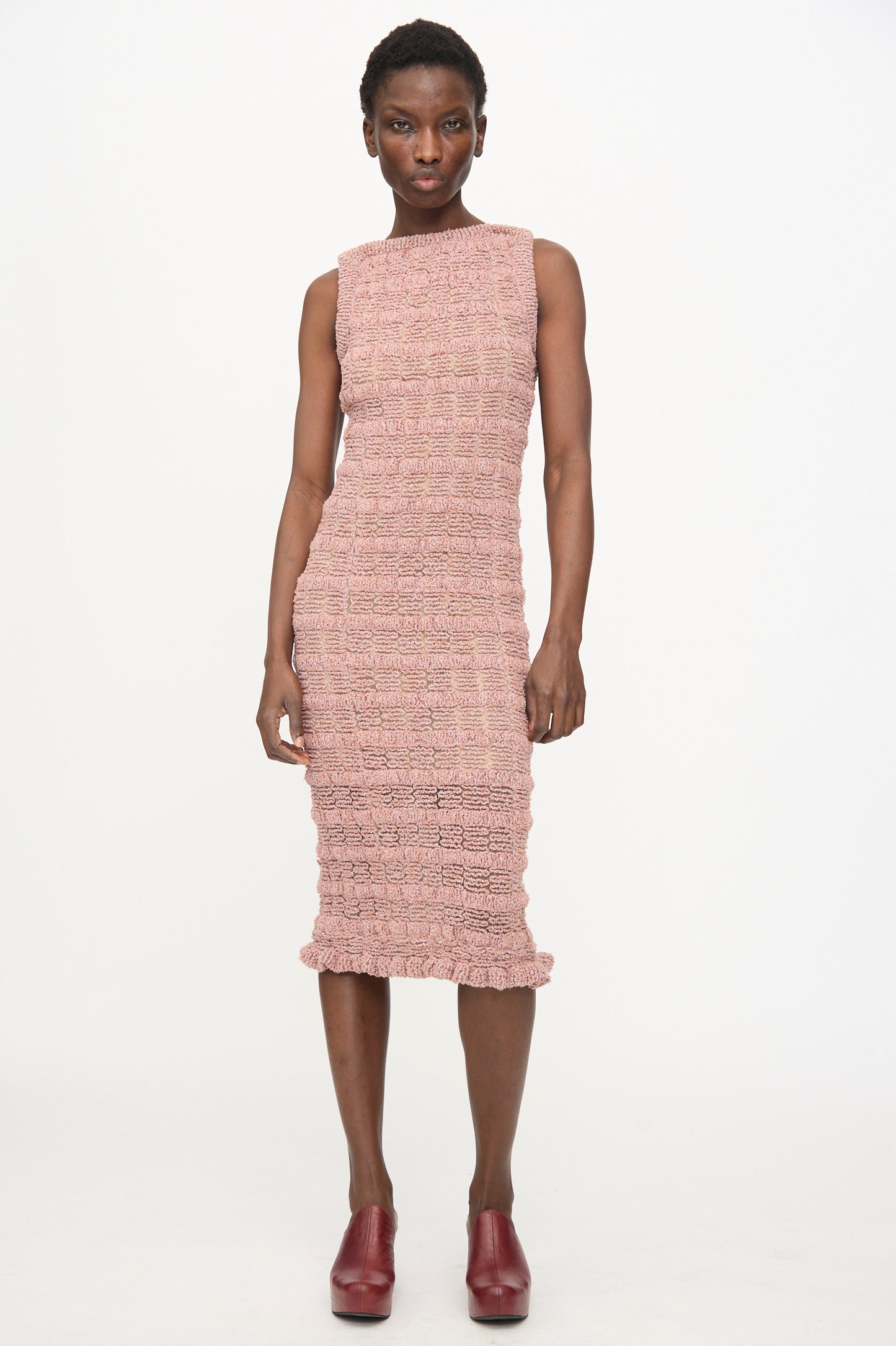 A person stands against a white background wearing the Veronique Leroy Smock Knit Dress in Dusk—a sleeveless, smocked midi dress in textured pink—paired with burgundy shoes.