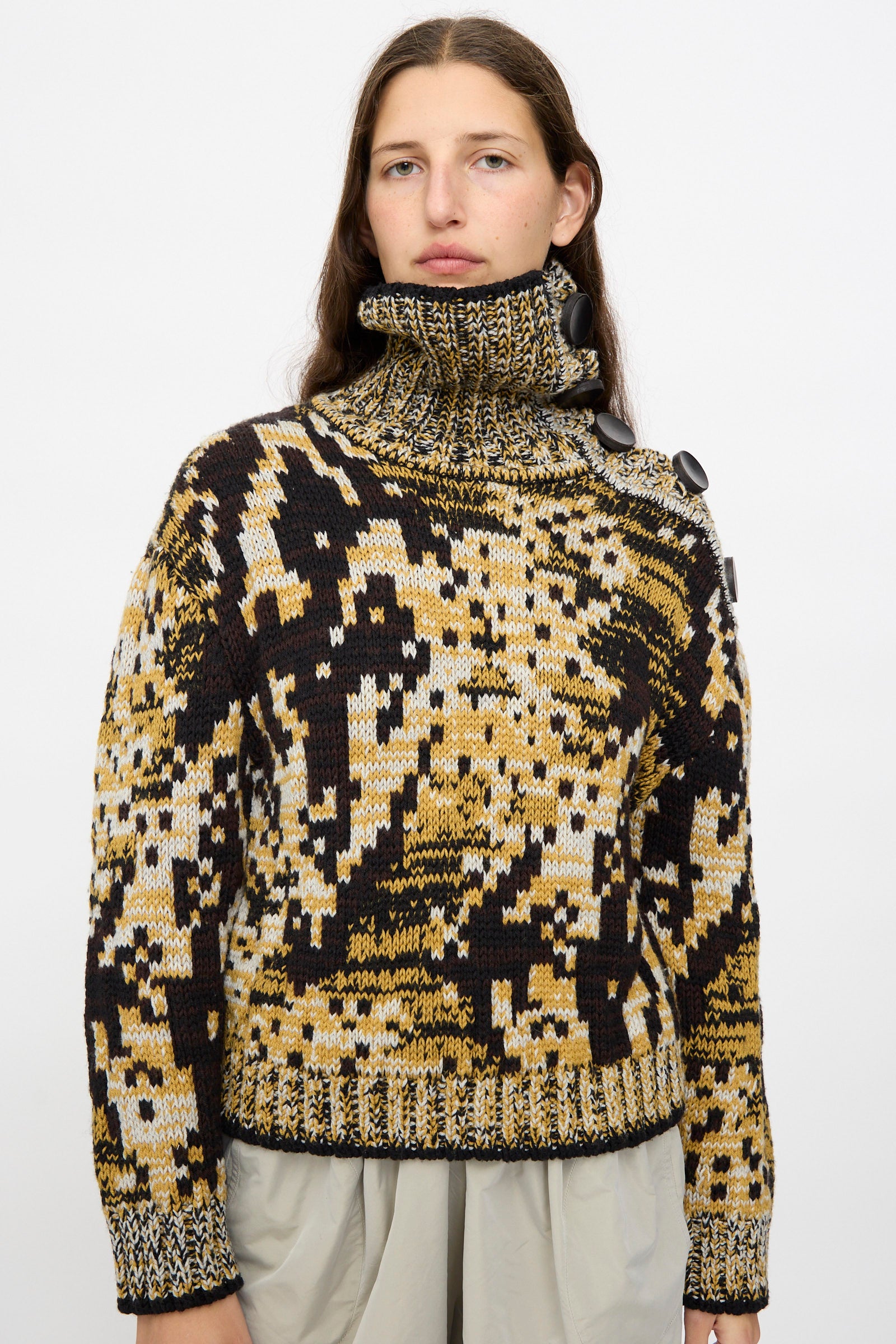 A person wears the Veronique Leroy Sweater in Sepia, a chunky high-neck knit with a yellow, black, and beige abstract pattern and large collar buttons, paired with light-colored pants.
