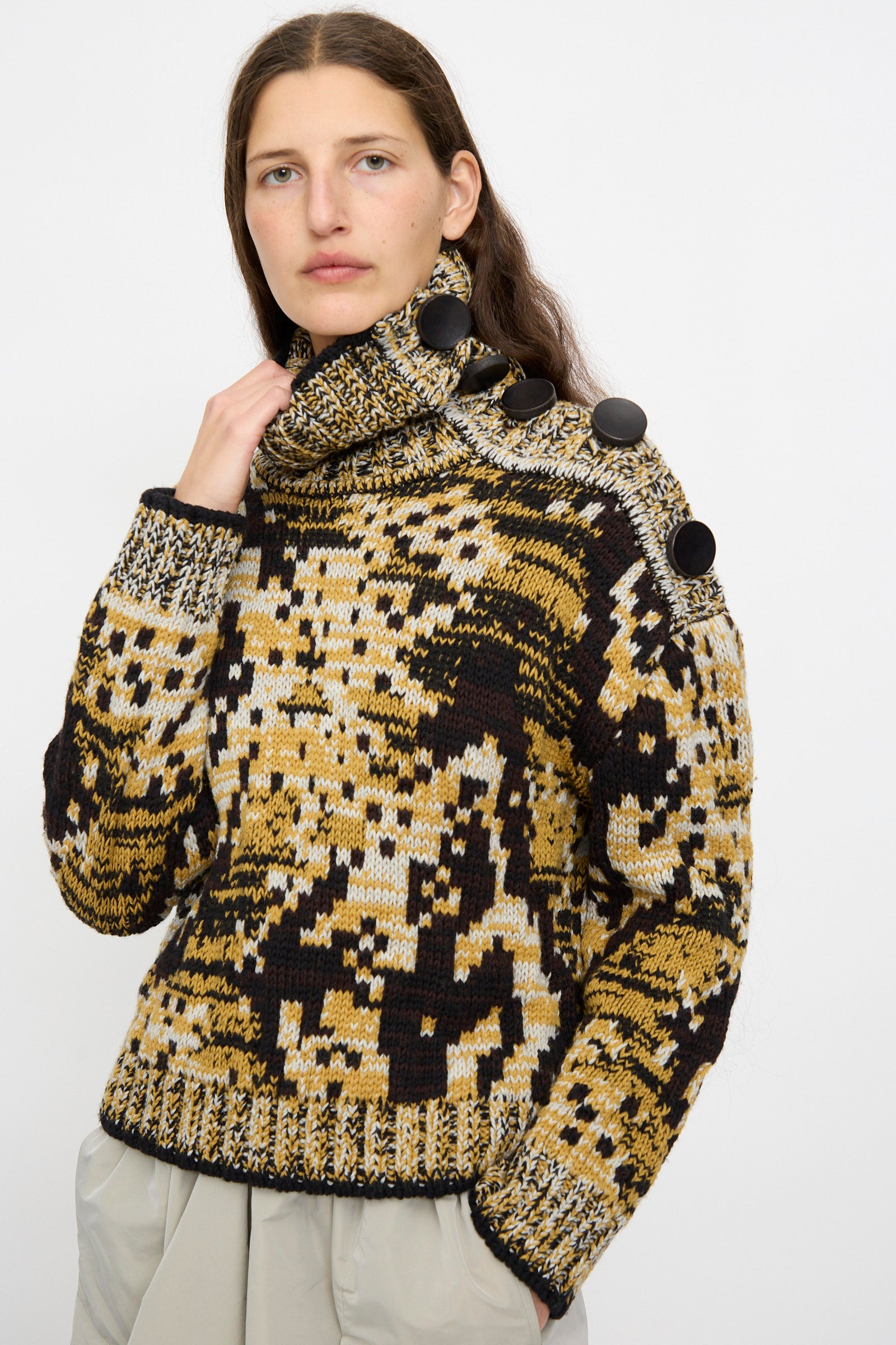 A person wears the Veronique Leroy Sweater in Sepia, featuring a patterned, high-neck relaxed fit with large shoulder buttons, paired with light-colored pants, standing against a plain background.