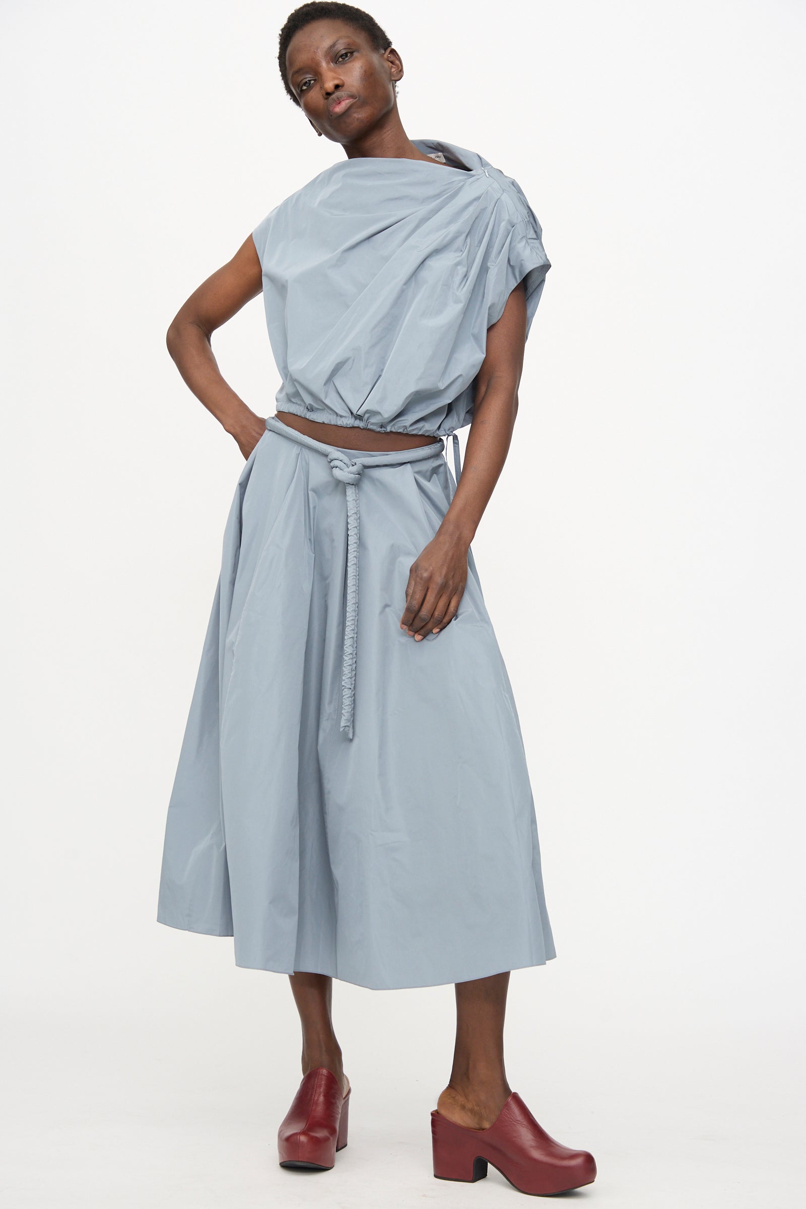 A model wears the Veronique Leroy Taffeta Skirt in Storm with a matching light blue sleeveless top, adjustable rope belt, and red platform clogs, posing against a plain white background.