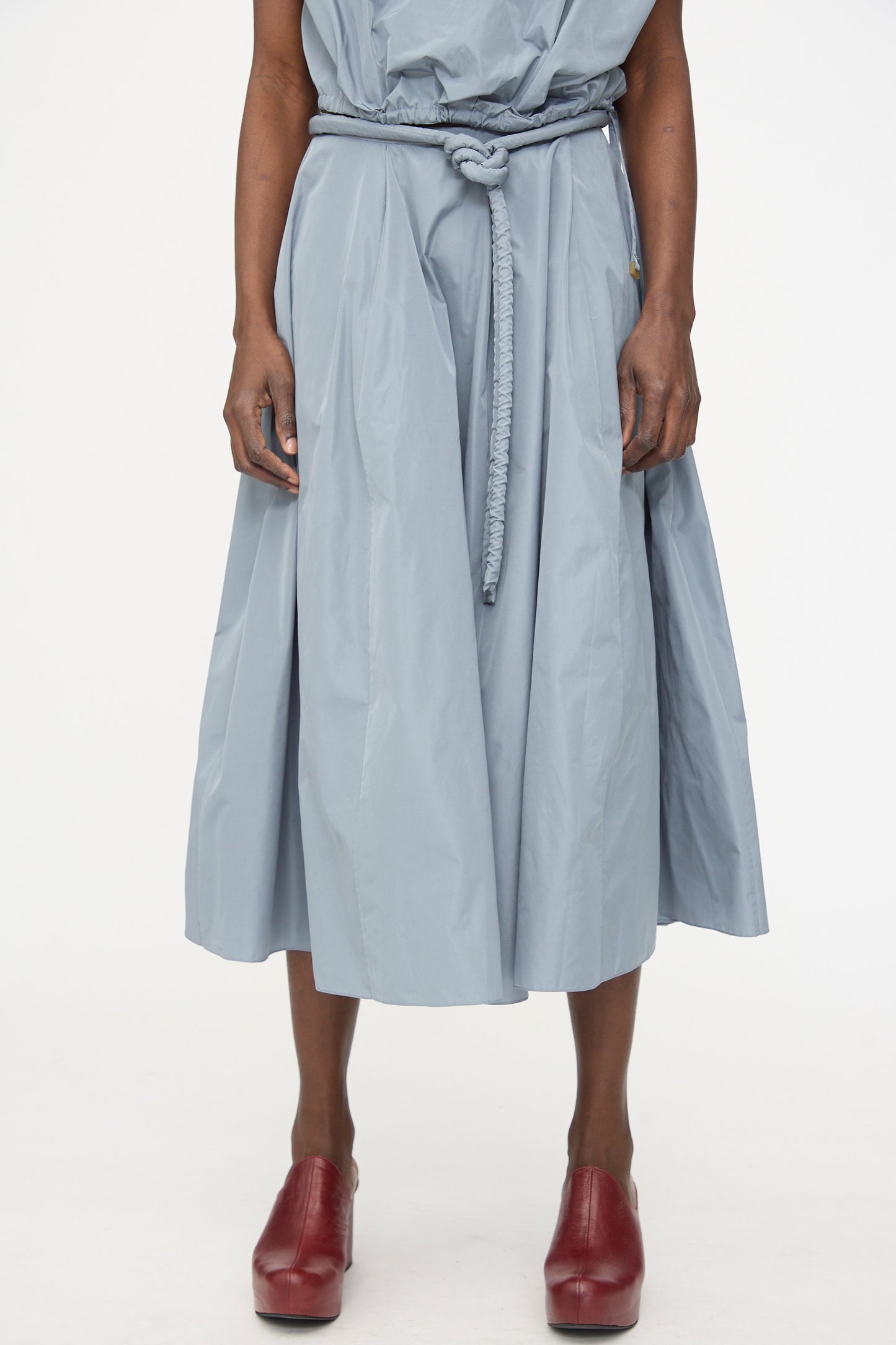 A person wears the Veronique Leroy Taffeta Skirt in Storm, styled with an adjustable rope belt and red clogs, featuring an A-line silhouette, standing against a plain white background.