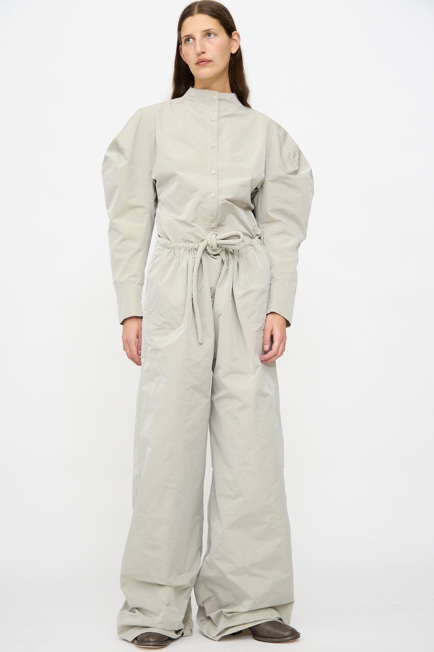 A woman wears Veronique Leroy's Tie Pants in Sage—a lightweight taffeta, high-waisted, wide-leg style with a button-up front and long sleeves—paired with brown shoes against a plain white background.