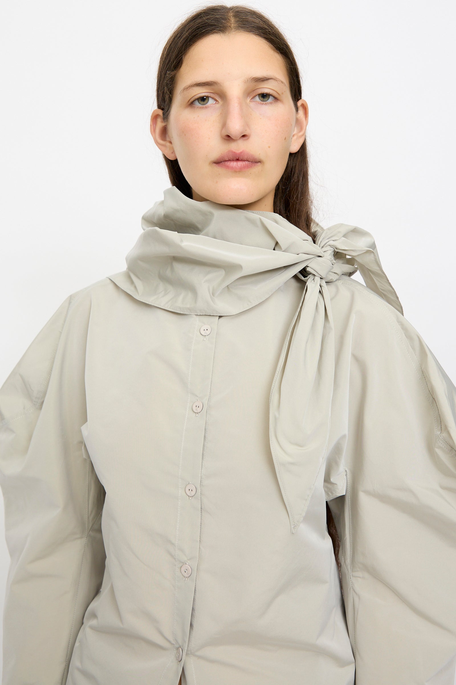 A woman with long brown hair models the Veronique Leroy Top with Scarf in Sage, featuring a large bow at the band collar, against a plain white background.