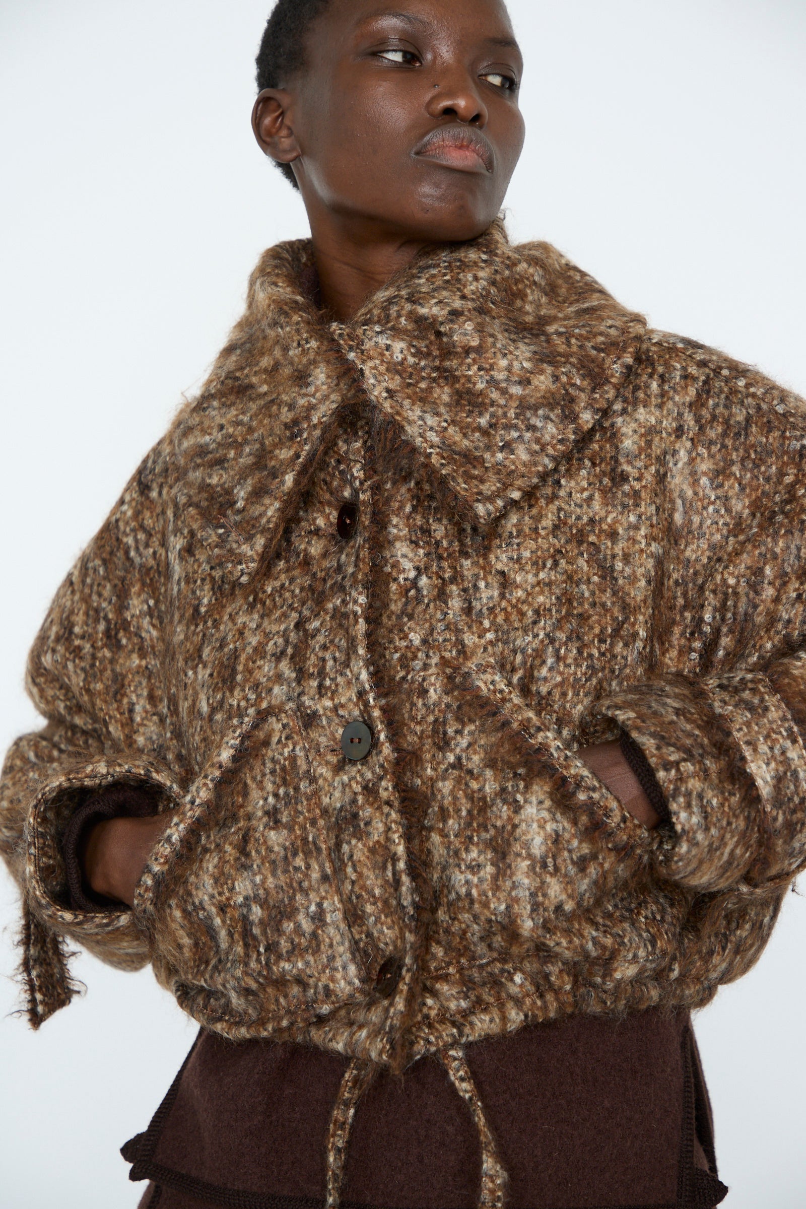 A person wearing the Tweed Bomber Jacket in Bronzewood by Veronique Leroy, made from virgin wool with a speckled brown pattern, large buttons, and an oversized collar, stands with hands in pockets against a plain background.