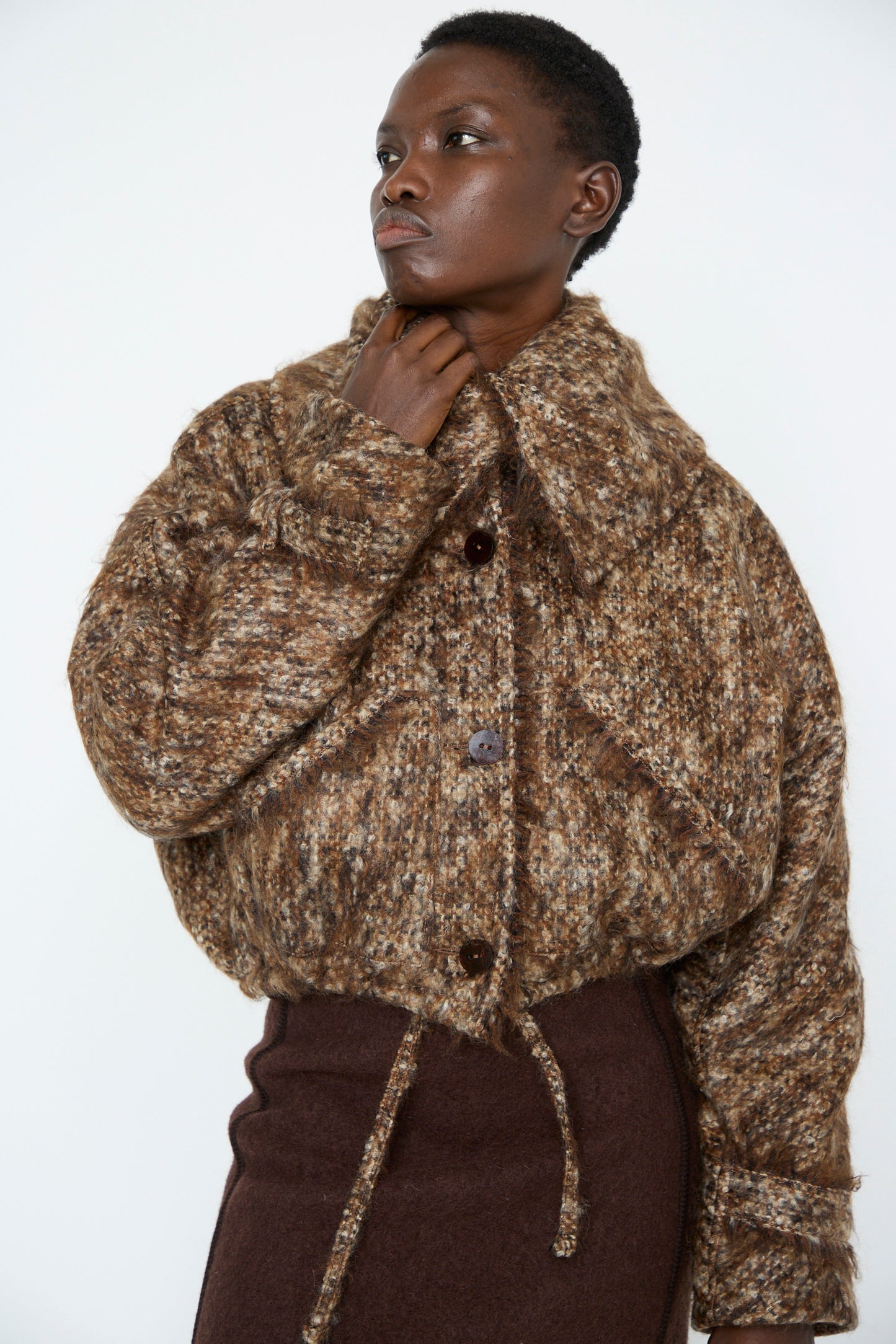 An individual wearing the Tweed Bomber Jacket in Bronzewood by Veronique Leroy, showing its virgin wool texture, large buttons, and oversized collar, paired with a brown skirt against a plain background.