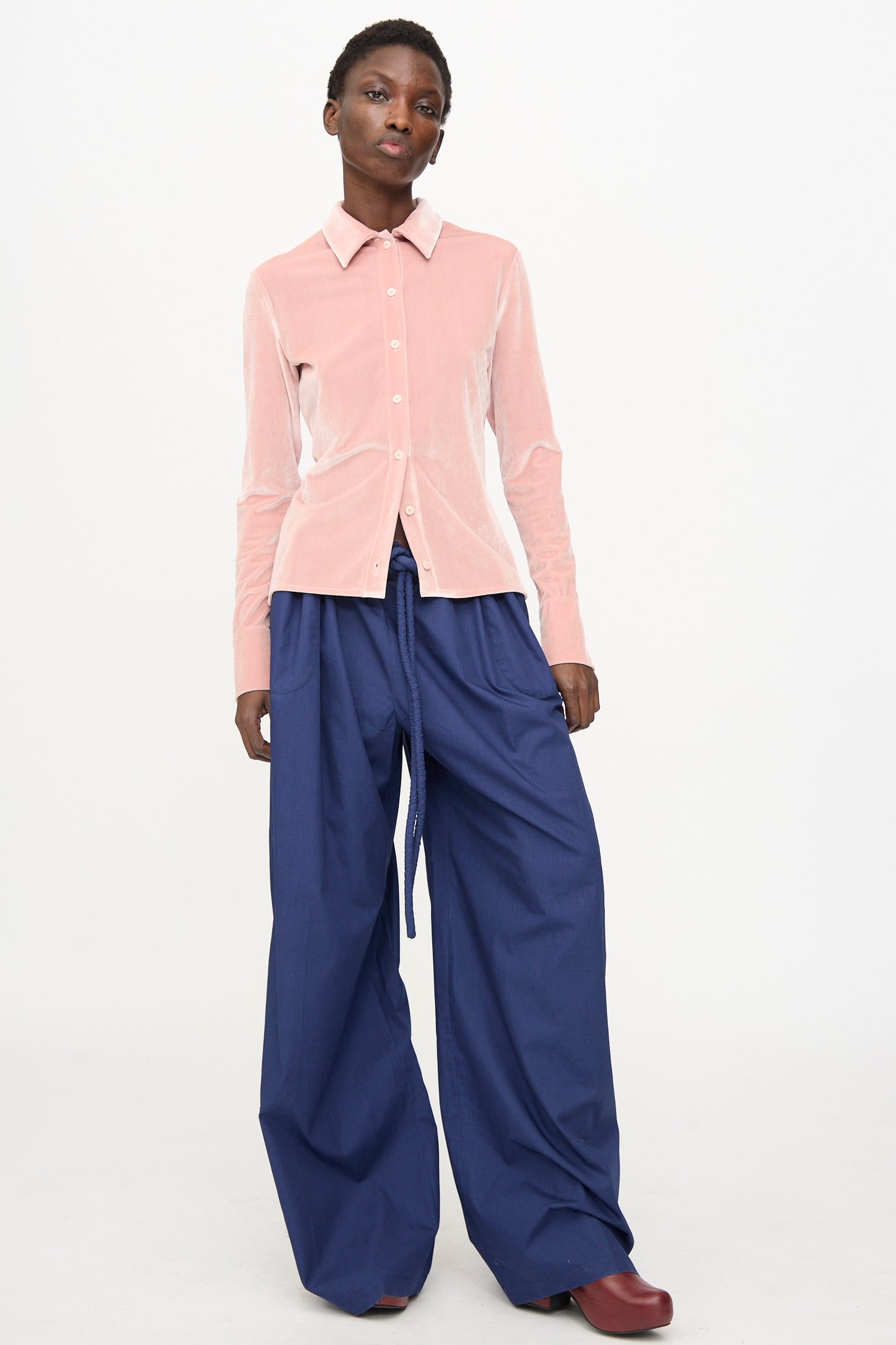 A person stands against a plain background wearing the Veronique Leroy Velvet Classic Shirt in Blush, paired with wide-leg blue drawstring pants and dark red shoes.