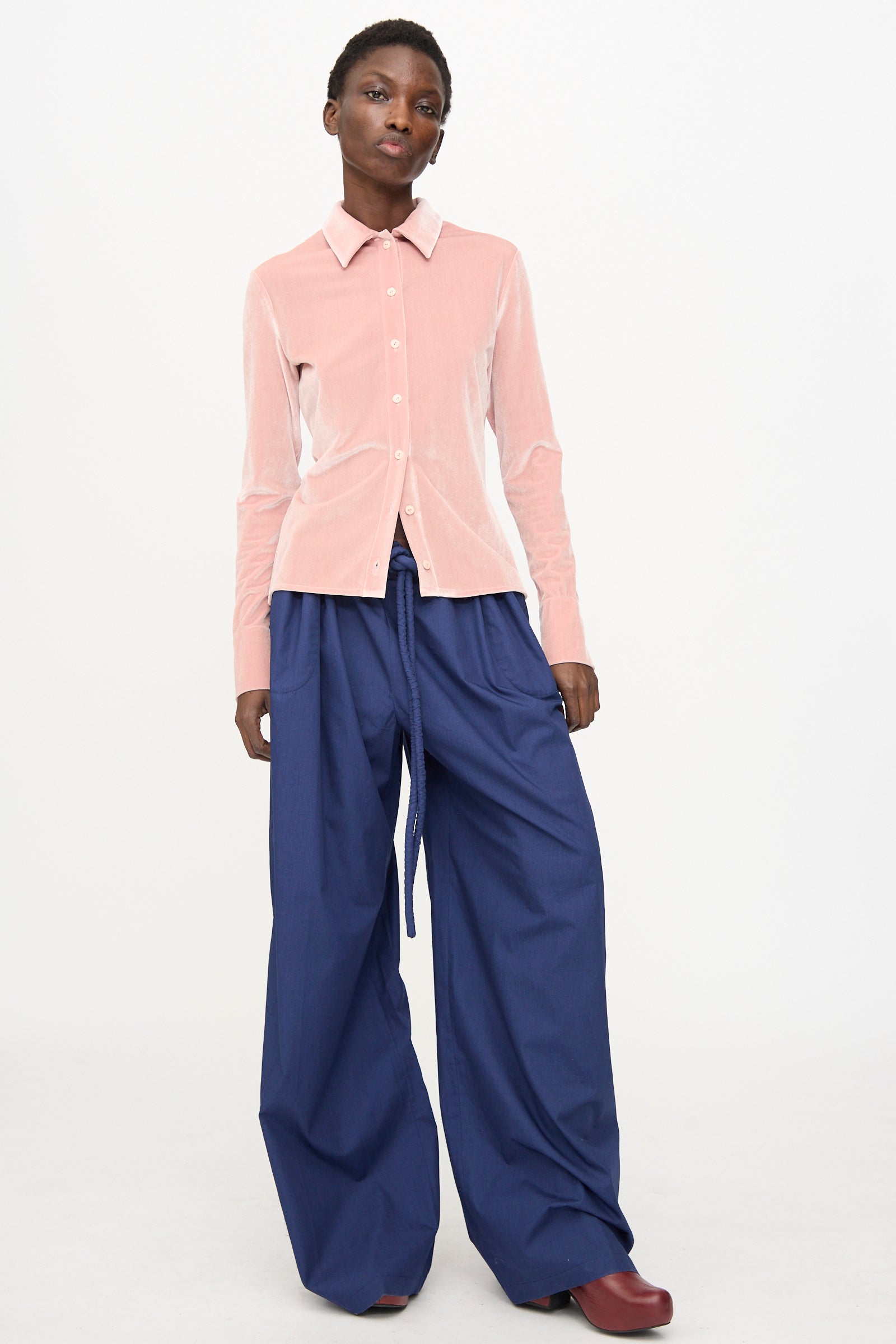 A person stands against a plain background wearing the Veronique Leroy Velvet Classic Shirt in Blush, paired with wide-leg blue drawstring pants and dark red shoes.