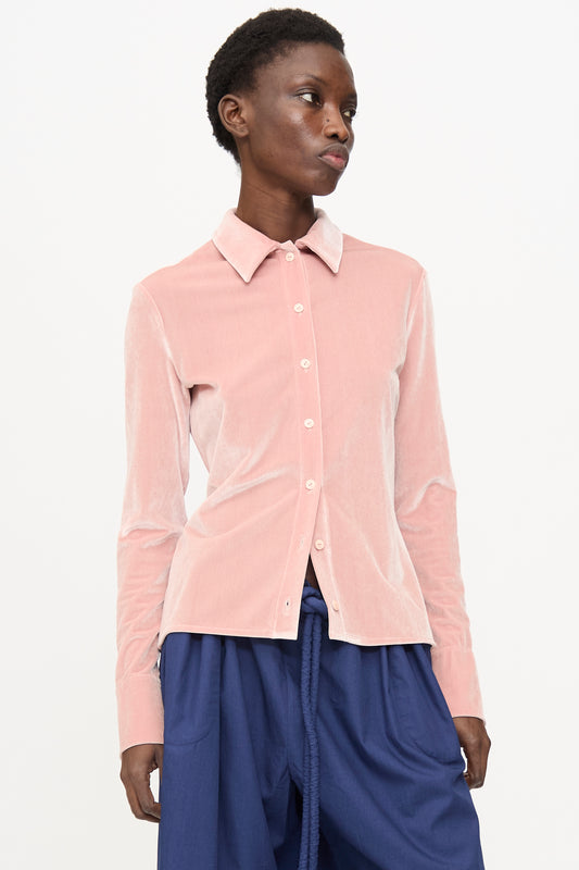 A person stands against a plain white background wearing the Veronique Leroy Velvet Classic Shirt in Blush, featuring a collared, long-sleeve design with buttons, paired with loose blue pants.