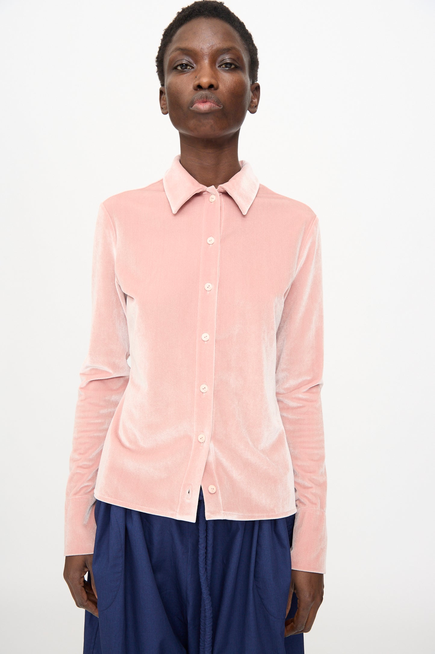 A person stands against a plain background, wearing the Veronique Leroy Velvet Classic Shirt in Blush—a relaxed-fit, collared, light pink button-up with long sleeves—paired with dark blue pants.