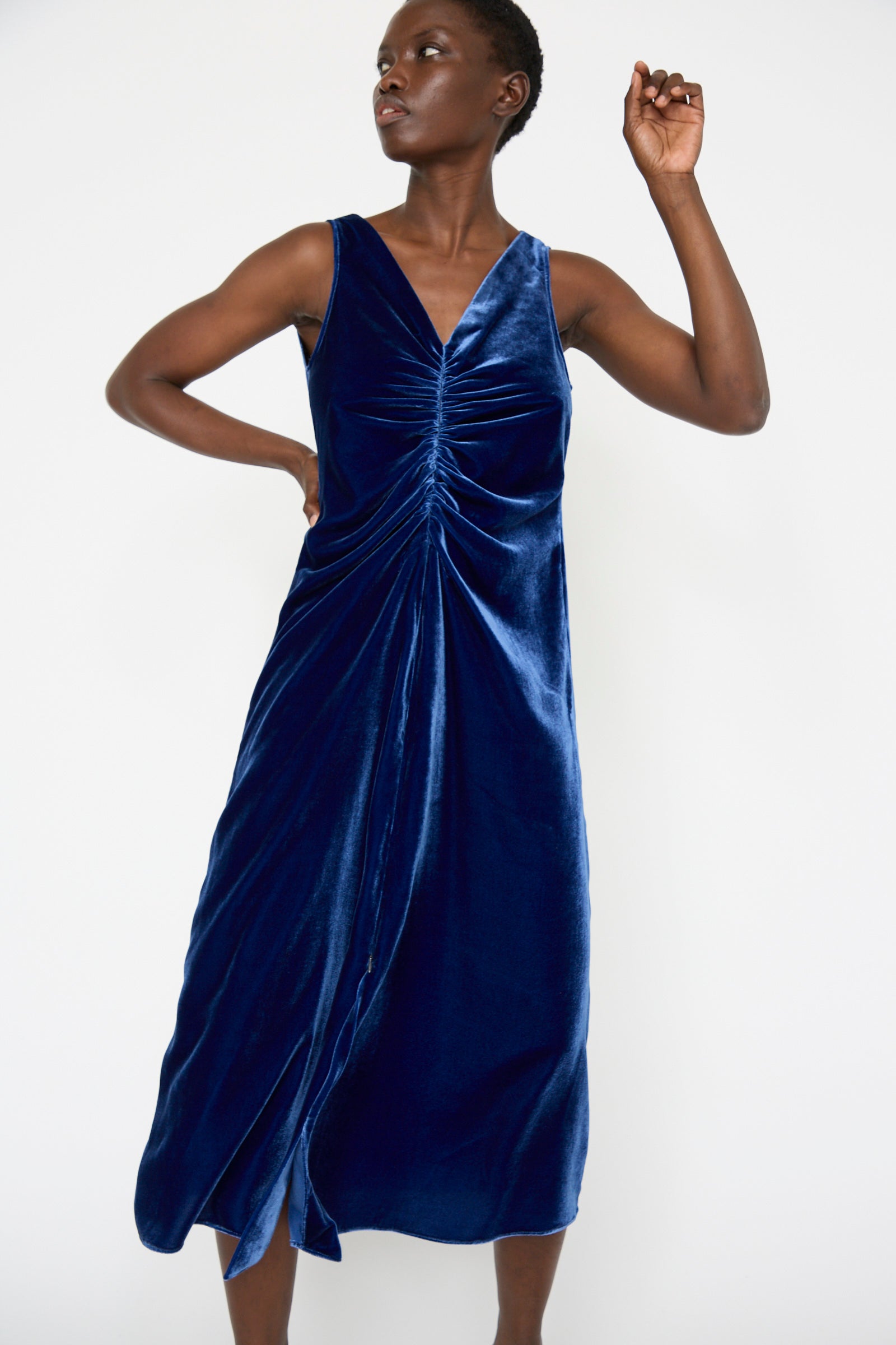 The woman stands against a plain white background, in Veronique Leroy's Velvet Gather Front Dress in Cobalt, featuring its sleeveless design with a ruched front and a v-neckline.