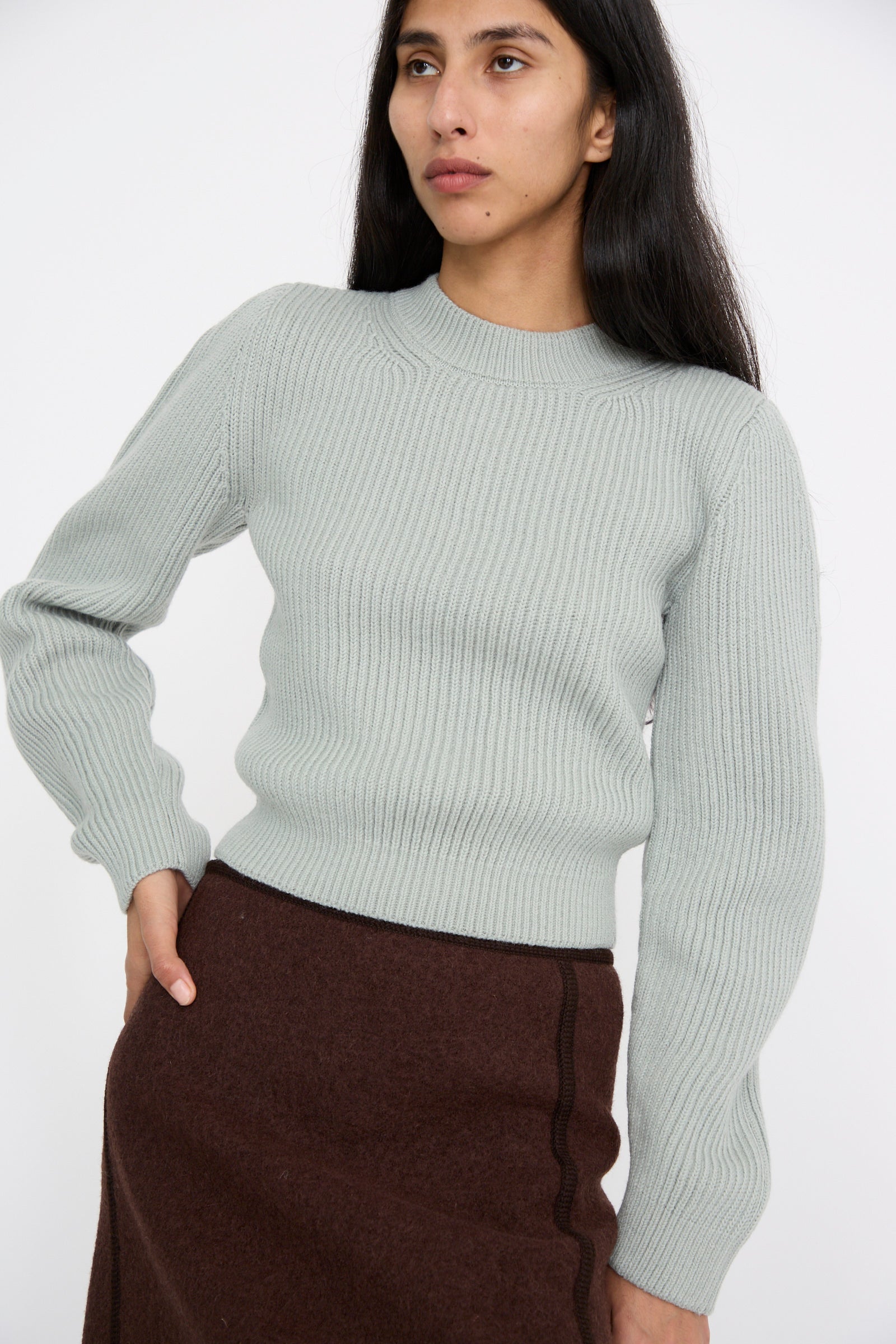 A person dressed in the Wavy Sleeve Sweater in Fog by Veronique Leroy and a brown skirt stands against a plain background with one hand on their hip.