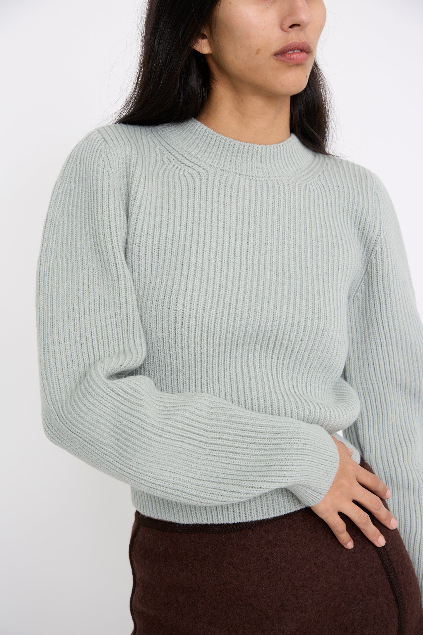 A person wearing the Wavy Sleeve Sweater in Fog by Veronique Leroy and a brown skirt stands against a plain white background with one hand on their right hip.