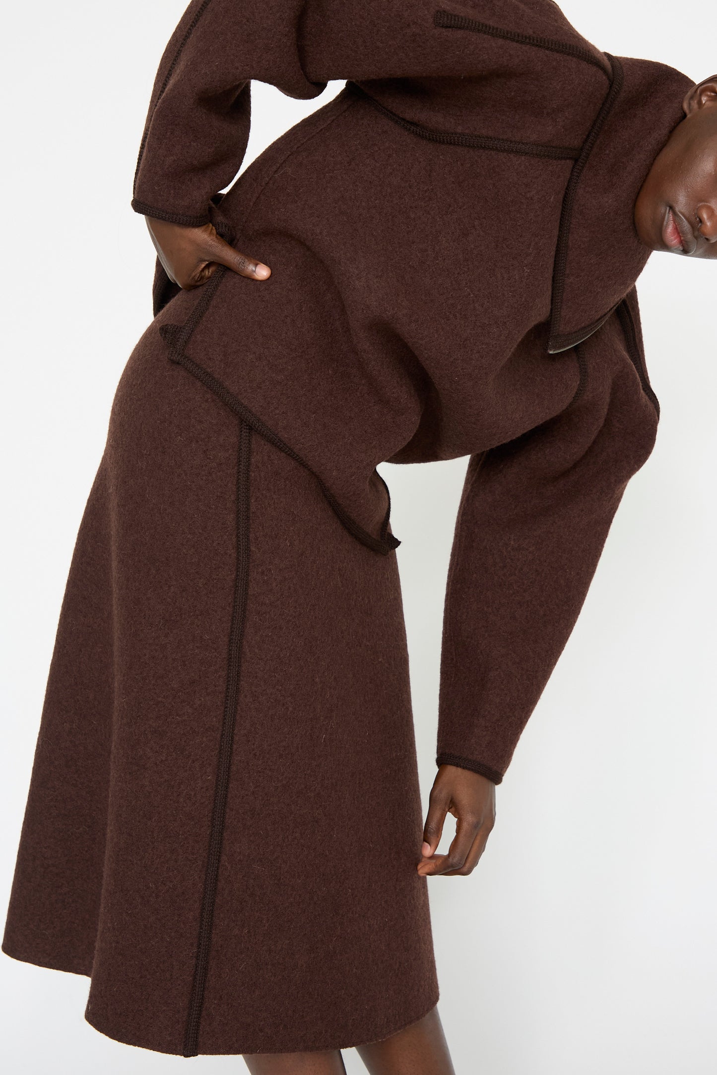 A person dressed in Veronique Leroy's Wool Long Skirt in Choco, accompanied by a coordinating long-sleeved top, bends slightly forward against a pristine white background.