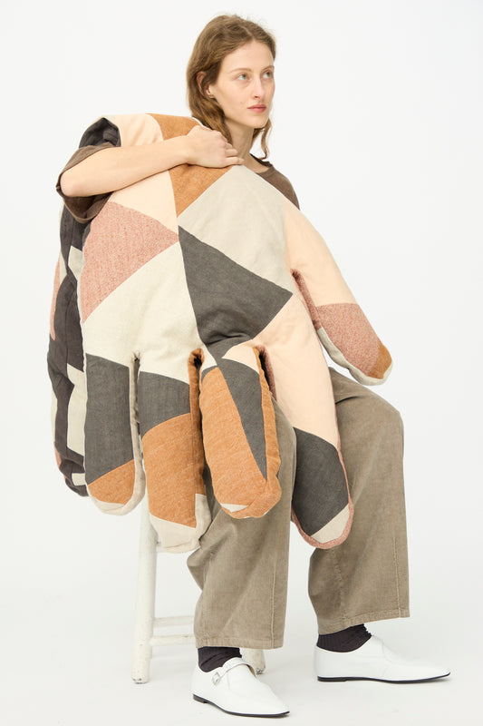 A person sits on a stool wearing the Sonia Delaunay Tissu Simultané Giant Hand Bolster I by Wiener Times—an oversized, patchwork quilted garment in neutral tones with wide pant legs and white shoes, set against a plain white background.