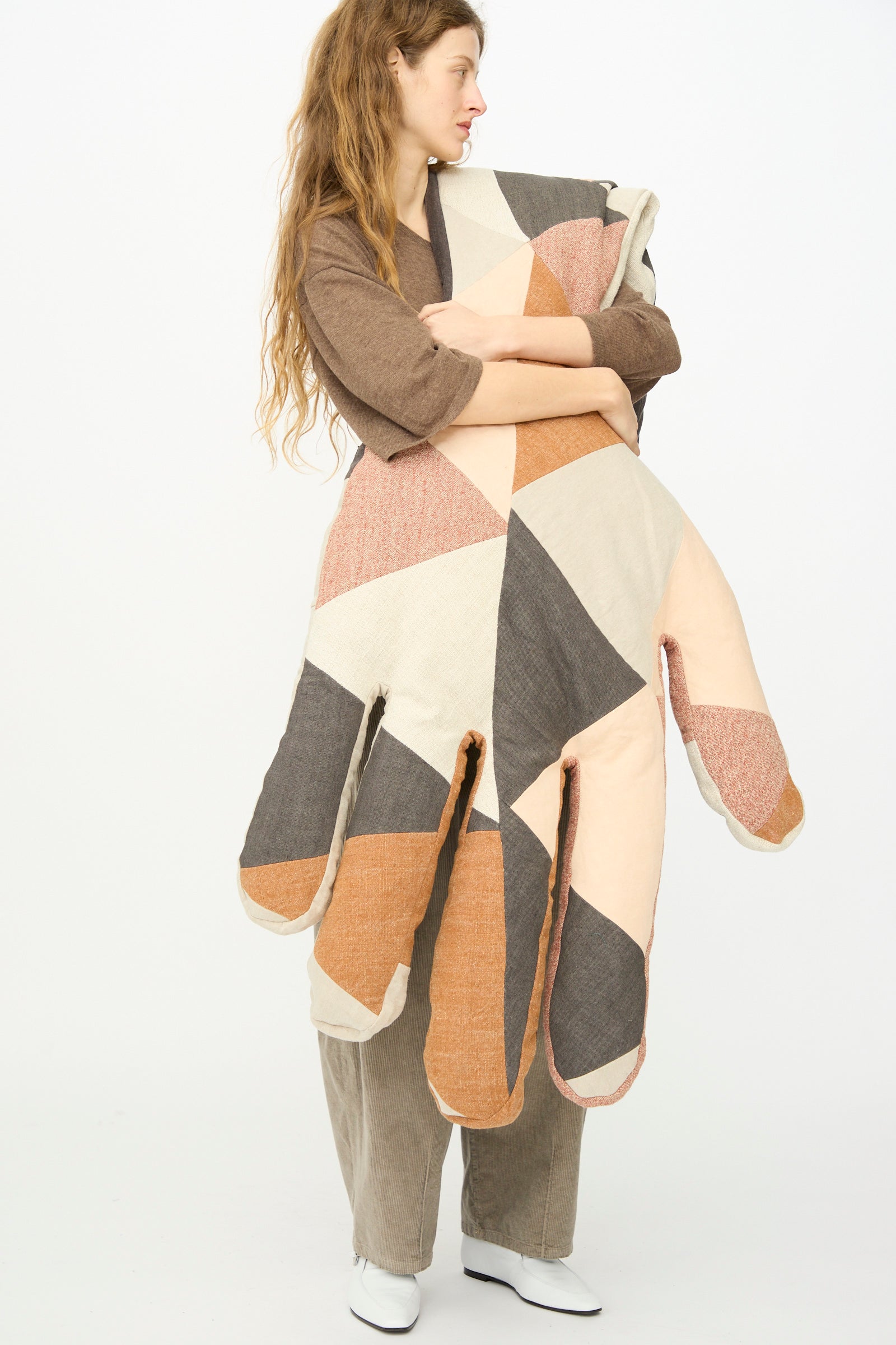 A person with long hair holds the Wiener Times Sonia Delaunay Tissu Simultané Giant Hand Bolster I, a large hand-shaped pillow made in Austria with beige, brown, orange, and black geometric patterns inspired by Wiener Werkstätte.