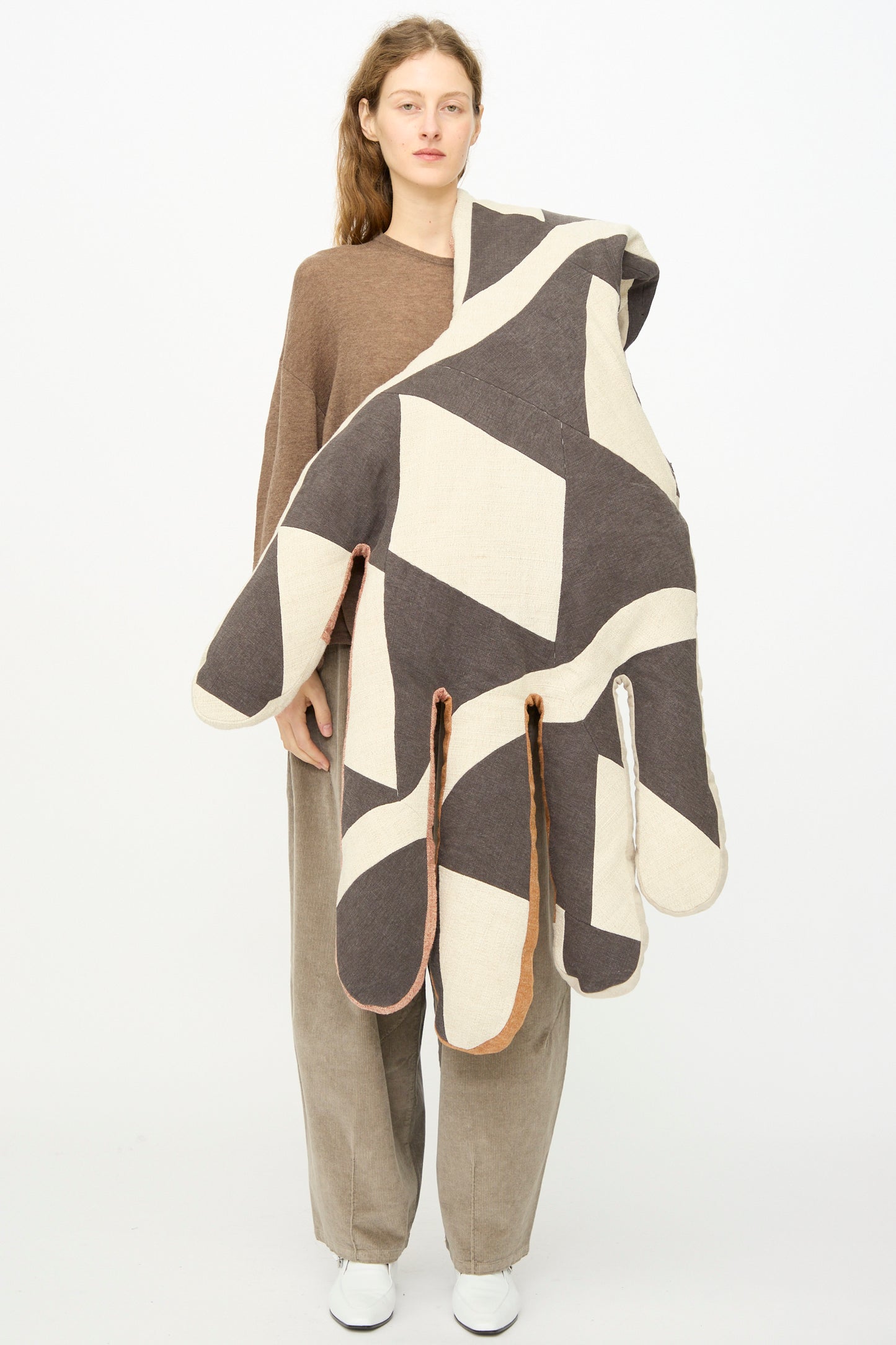 A woman faces forward in neutral-toned clothing, holding the Sonia Delaunay Tissu Simultané Giant Hand Bolster I by Wiener Times, a large hand-shaped pillow inspired by the Wiener Werkstätte.